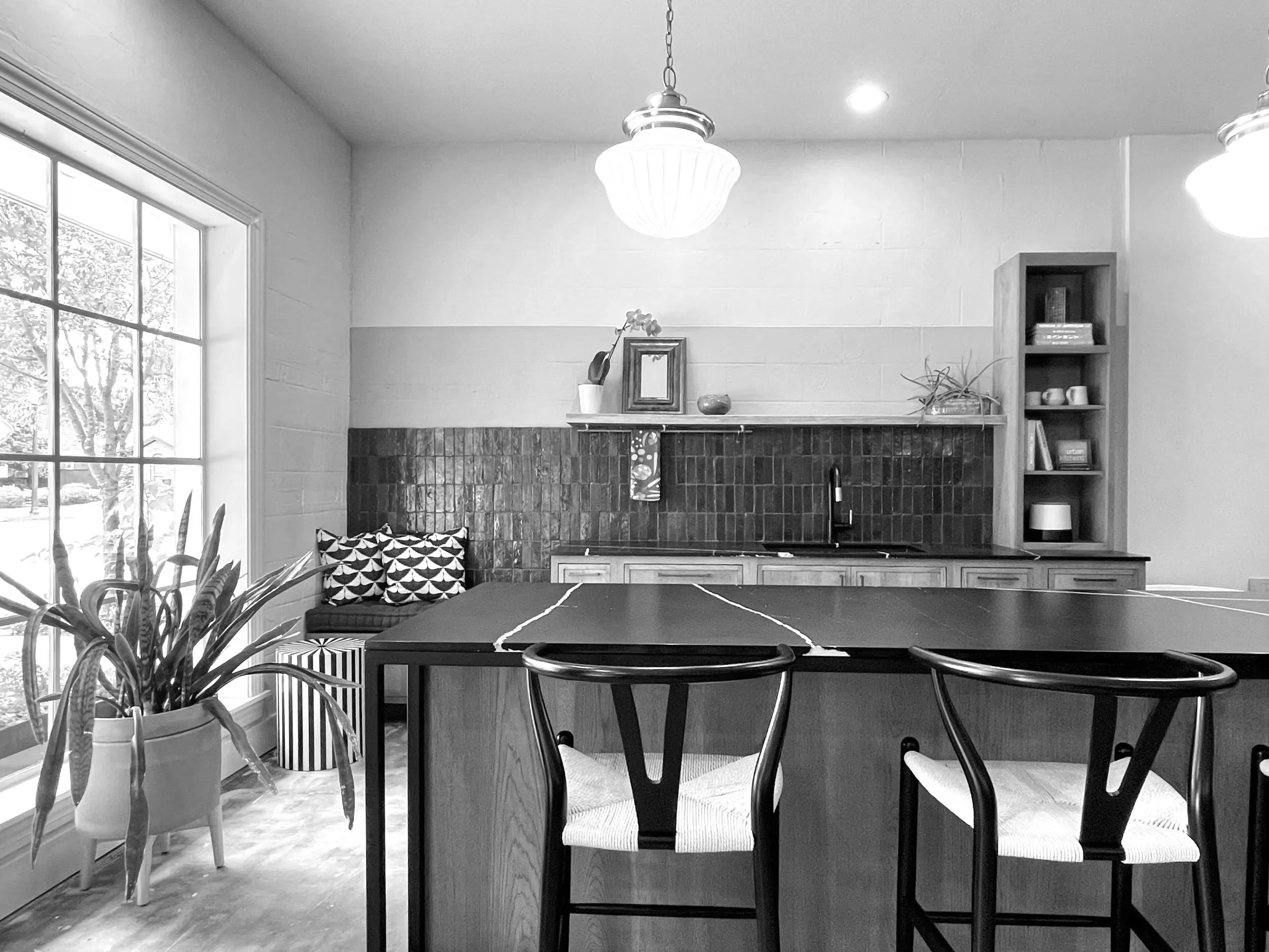 A dining area with a wooden table and black chairs with woven seats. A large window with grid panes lets in natural light. A potted plant with striped leaves is on the left. In the background, a brick fireplace has a mantle with decorative items including a mirror, a vase, and flowers. A bookshelf on the right contains books and cups. Ceiling lights illuminate the room.