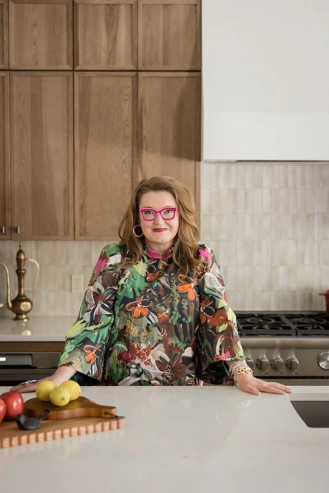 A woman with red hair, pink glasses, and floral clothing standing in a modern kitchen.