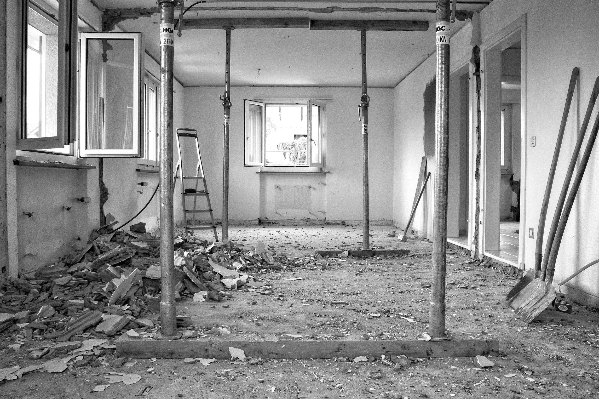 An interior space under renovation with construction debris on the floor, exposed support poles, and open window.