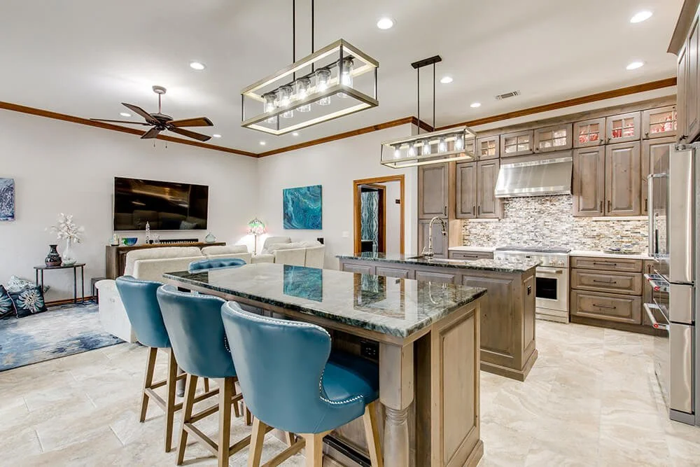 Open-concept kitchen with gray cabinetry, granite island, stainless steel appliances, tile backsplash, and blue and beige decor in the adjacent living room.