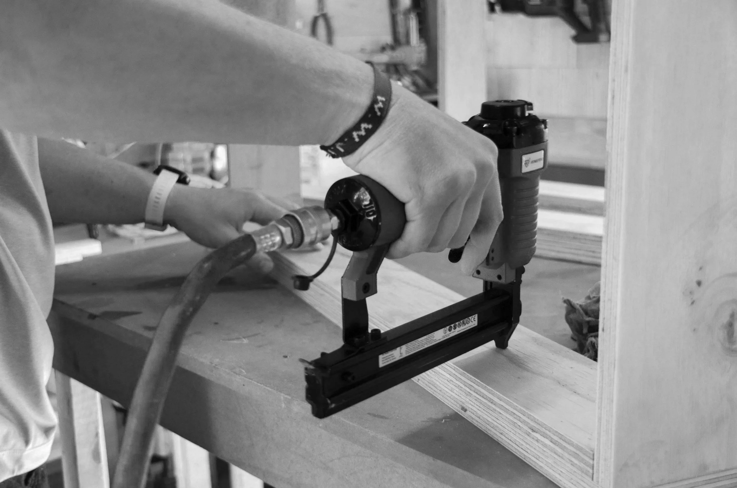A person operating a pneumatic nail gun to attach a piece of wood to a large wooden board in a woodworking shop.