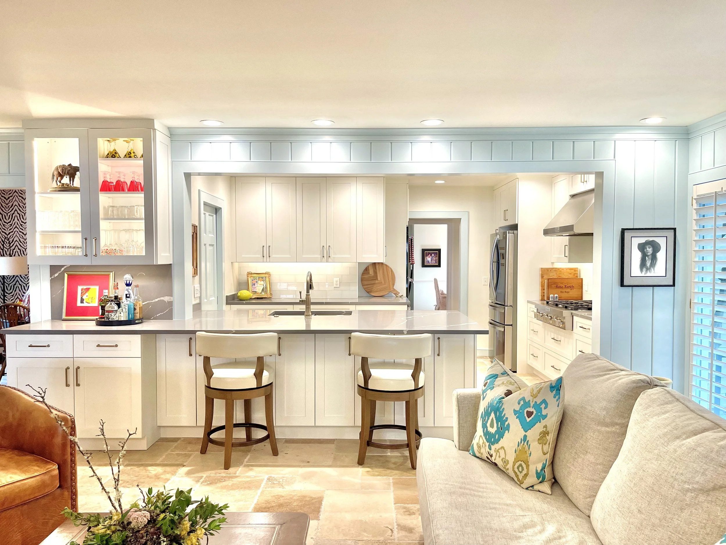 Open-concept kitchen and living room with cream cabinetry, stainless steel appliances, a kitchen island with beige bar stools, a beige sofa with decorative pillows, and a patterned armchair. Recessed lighting and decorative items are visible.