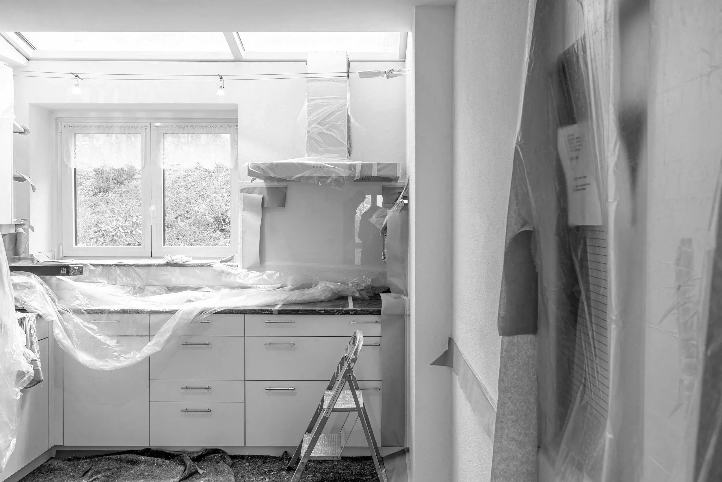 A kitchen undergoing renovation with a ladder, protective plastic coverings, and newly installed cabinets and countertops. There are window and ceiling lights visible.