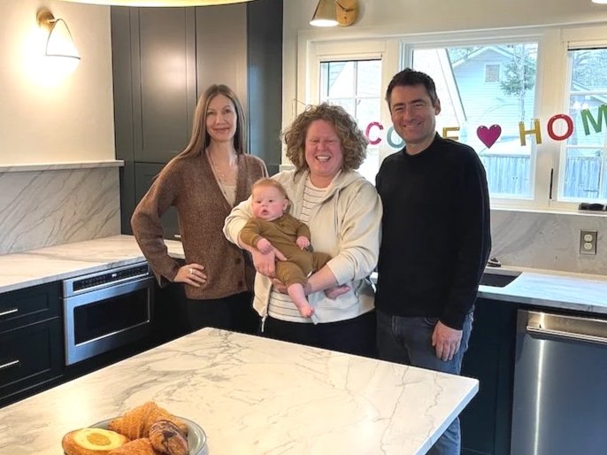 Four people, including a baby, standing in a kitchen with a marble island, smiling at the camera, with a 'Welcome Home' banner in the window.