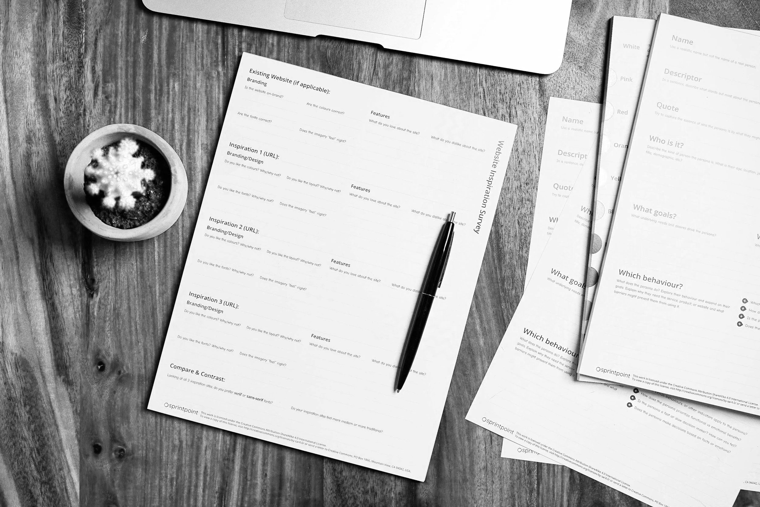 Black and white photograph of printed worksheets on a rustic wooden table. The worksheets include sections for website analysis and branding, with questions about site features, design, and inspiration. A black pen is placed on one of the sheets. To the left, there is a small white bowl containing a latte with a foam heart design. Part of a closed laptop is visible at the top of the image.