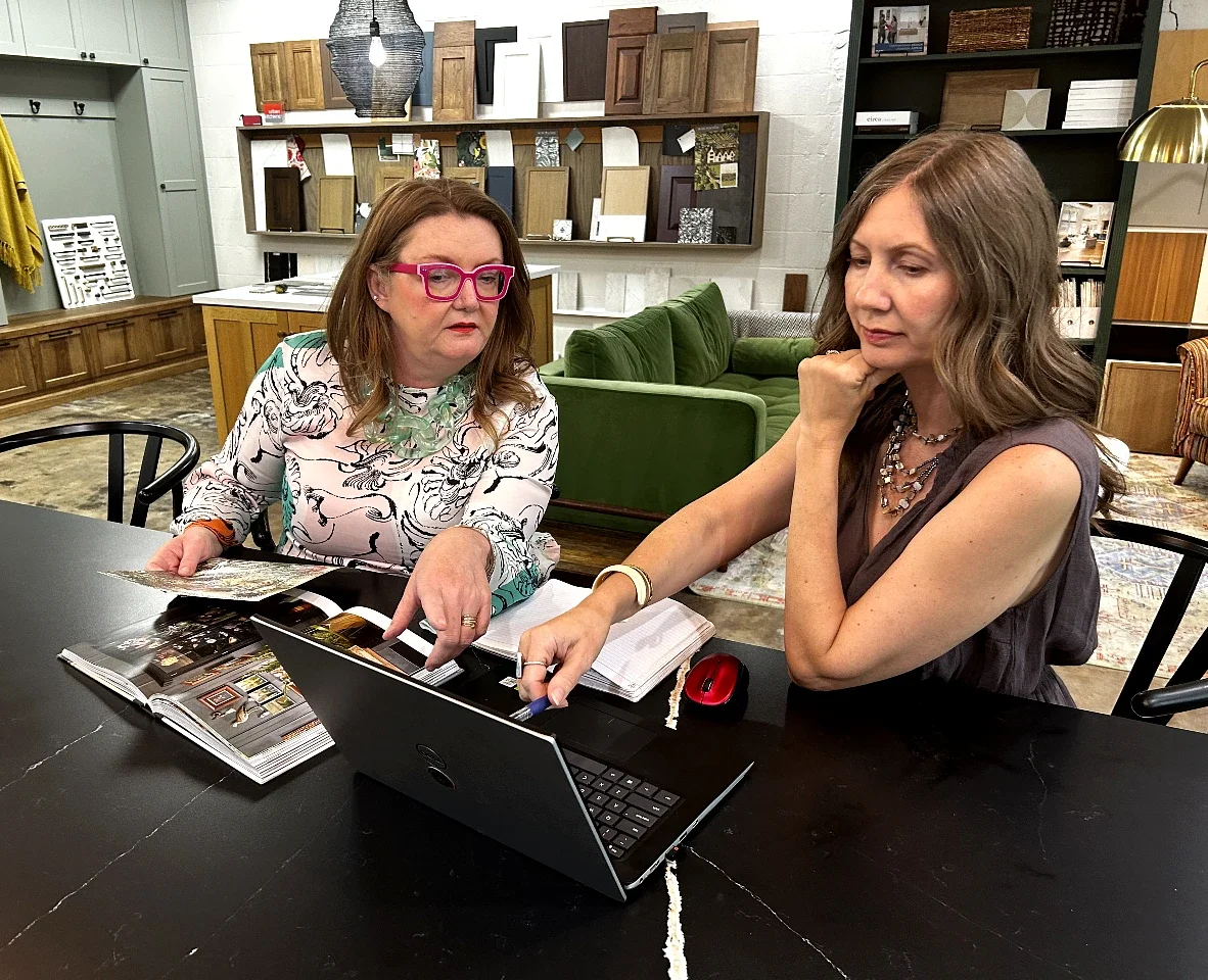 Two women are sitting at a black table, looking at a laptop and discussing. They have notebooks and magazines in front of them. The background shows shelves with decorative items, wood samples, and a green couch.