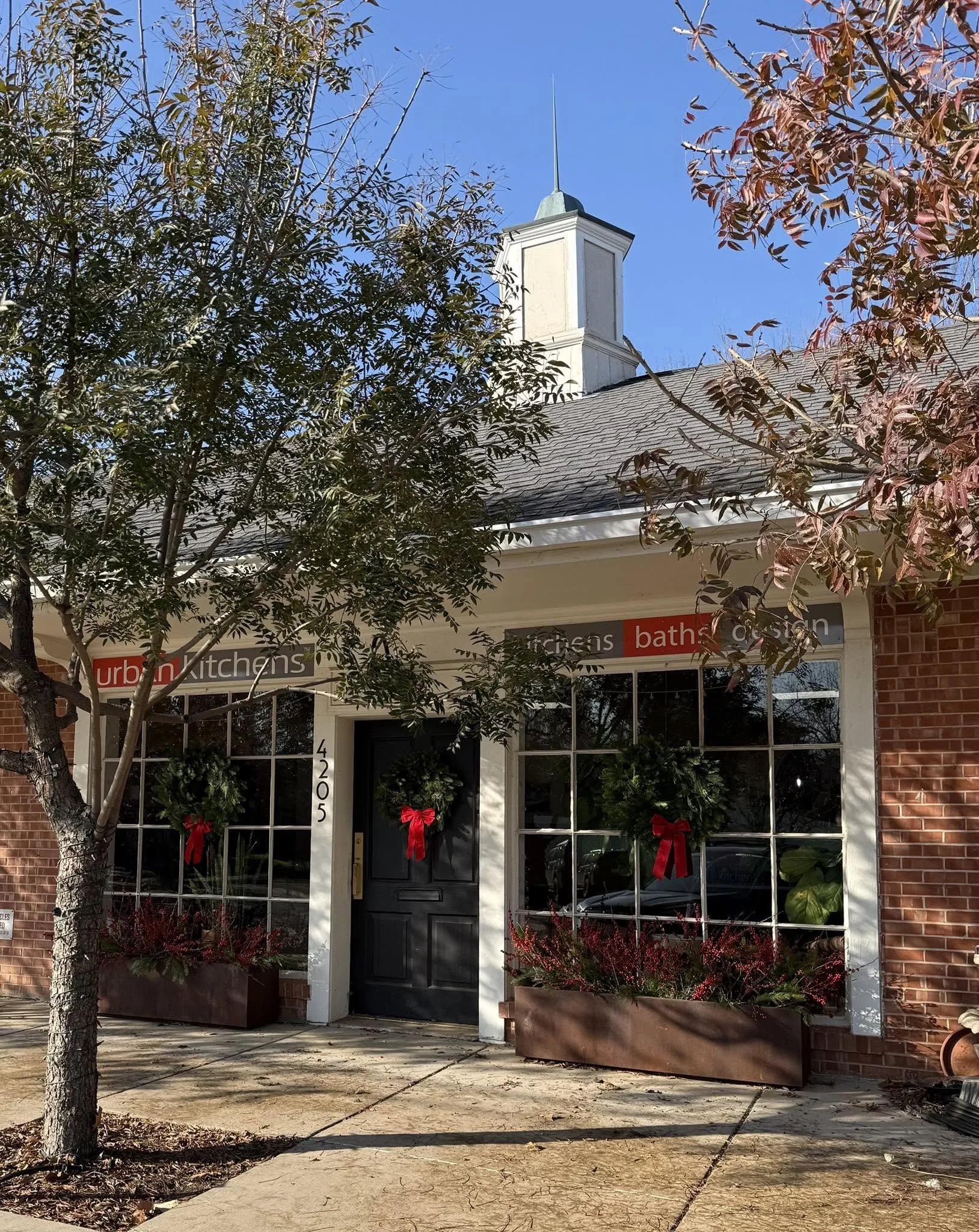 Storefront with holiday decorations, including wreaths with red ribbons, flanked by planters with red berries, two trees with some red leaves, and a sign reading 'urban kitchens' and 'bath' in the window, with a white building and blue sky in the background.