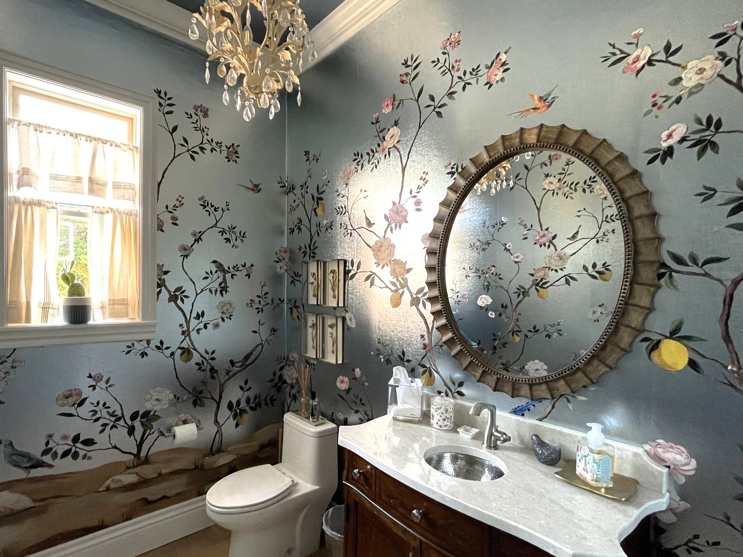 A bathroom with walls painted light blue, featuring a mural of pink and white flowers with green leaves and hummingbirds. There is a round mirror with a decorative frame, a window with light curtains, a chandelier hanging from the ceiling, a white countertop with a sink, and various toiletries and decorative items.