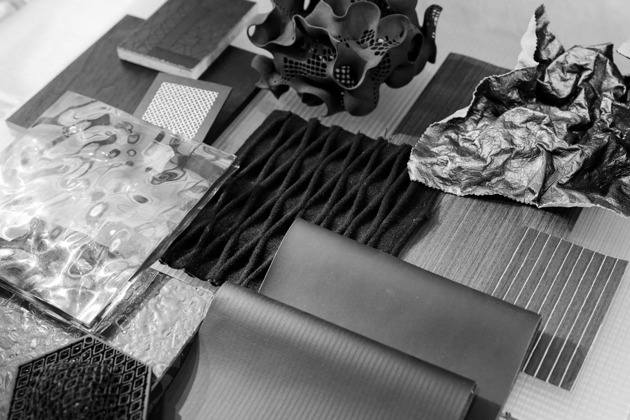 A collection of textured materials and objects on a wooden table, including crumpled paper, a plastic bag with water droplets, rhinestones, a ribbed fabric swatch, and a decorative geometric object.
