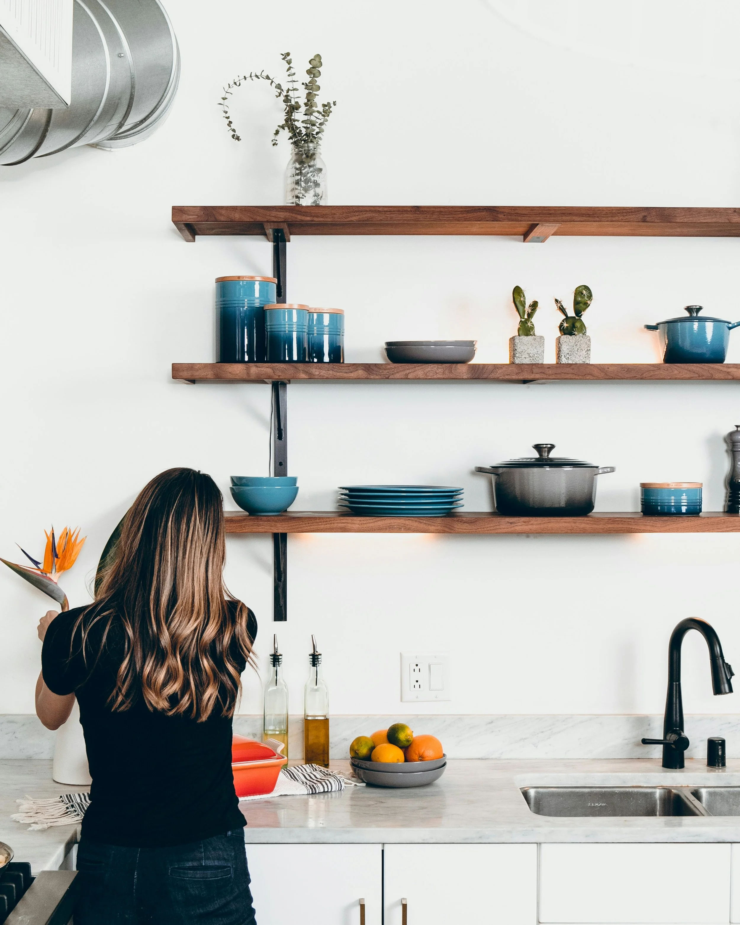 A woman with long wavy hair wearing a black shirt and jeans, standing in a modern kitchen, holding a glass with a flower in her left hand and facing away from the camera. The kitchen has wooden open shelves with blue dishes, a jar with dried eucalyptus, cacti in concrete pots, a bowl of fruit, and some oil bottles on the white marble countertop. A stovetop and a sink are also visible.