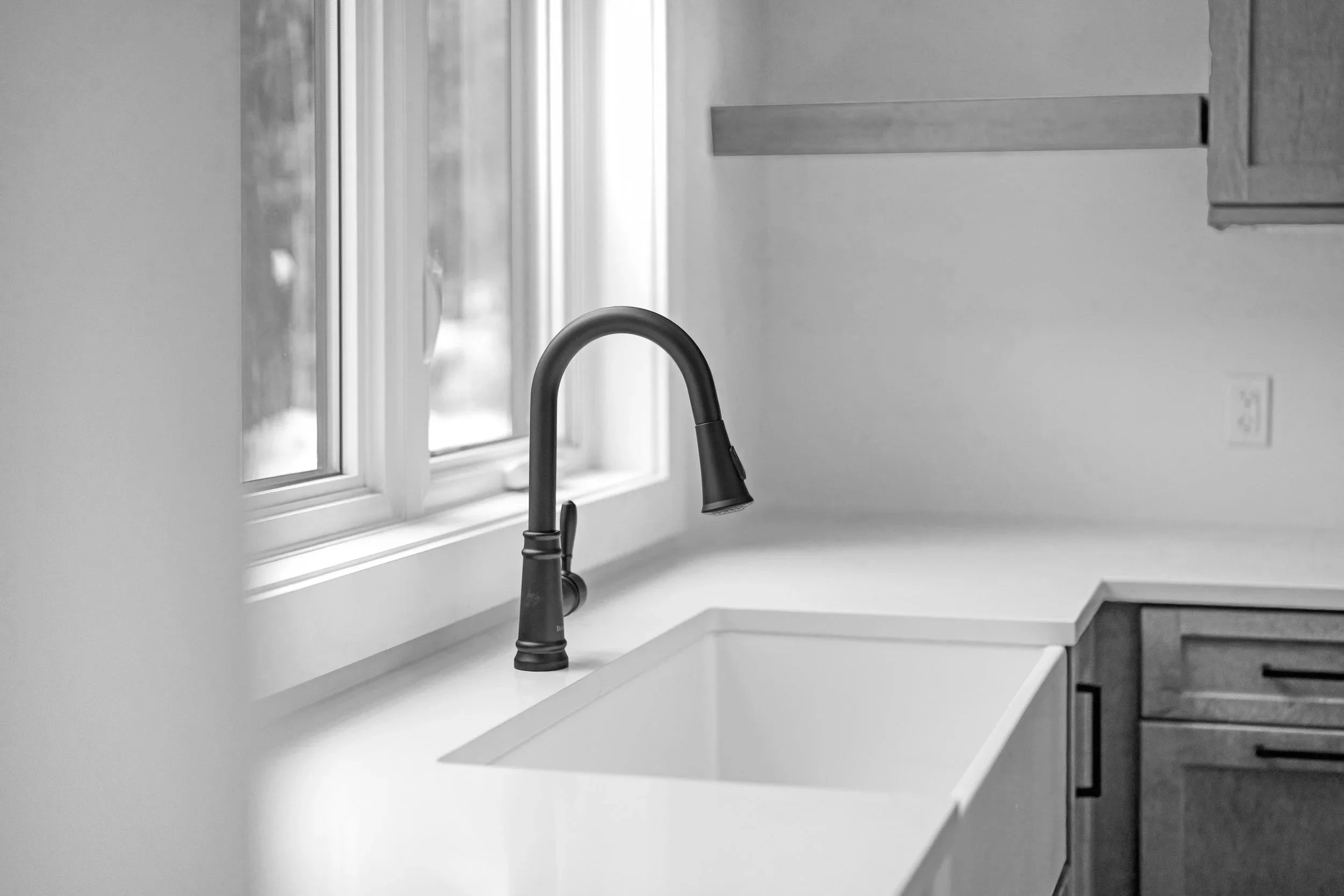 A kitchen sink with a black gooseneck faucet, situated below a window with white trim, and wooden cabinets above and below the countertop.