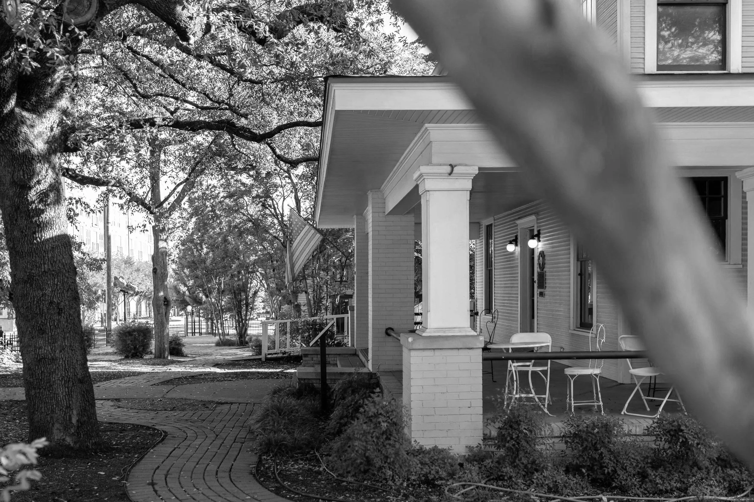 A brick and wood porch of a house with outdoor seating and an American flag, surrounded by trees and a brick pathway.