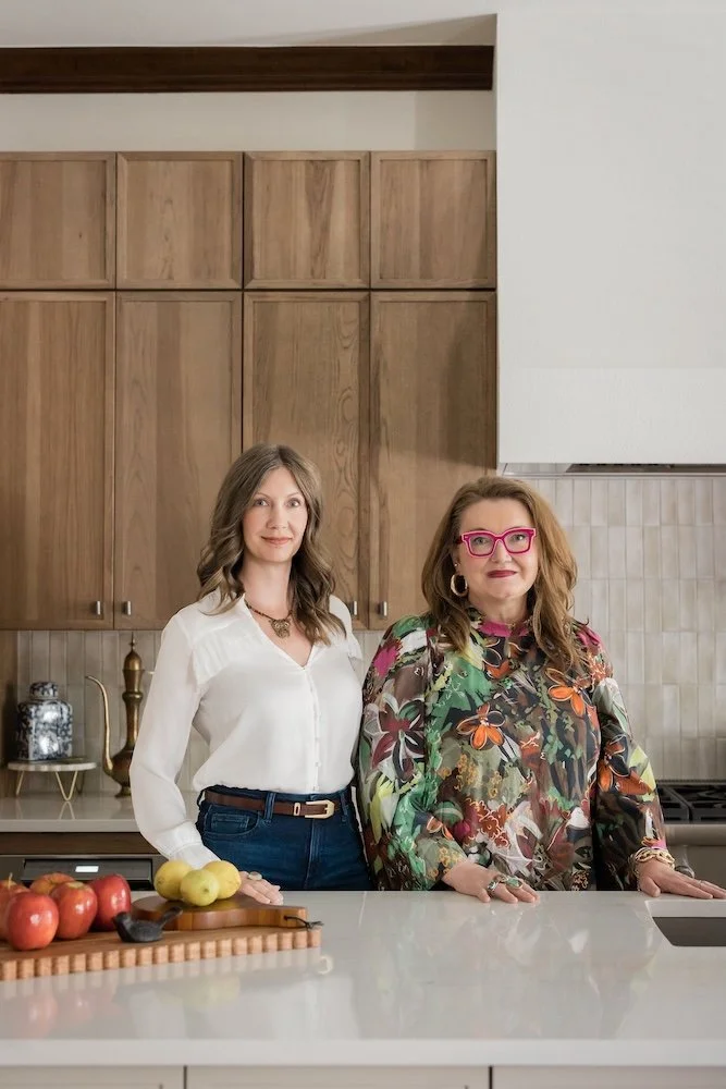 Two women standing in a modern kitchen behind a white countertop with apples and yellow fruit in front of them.