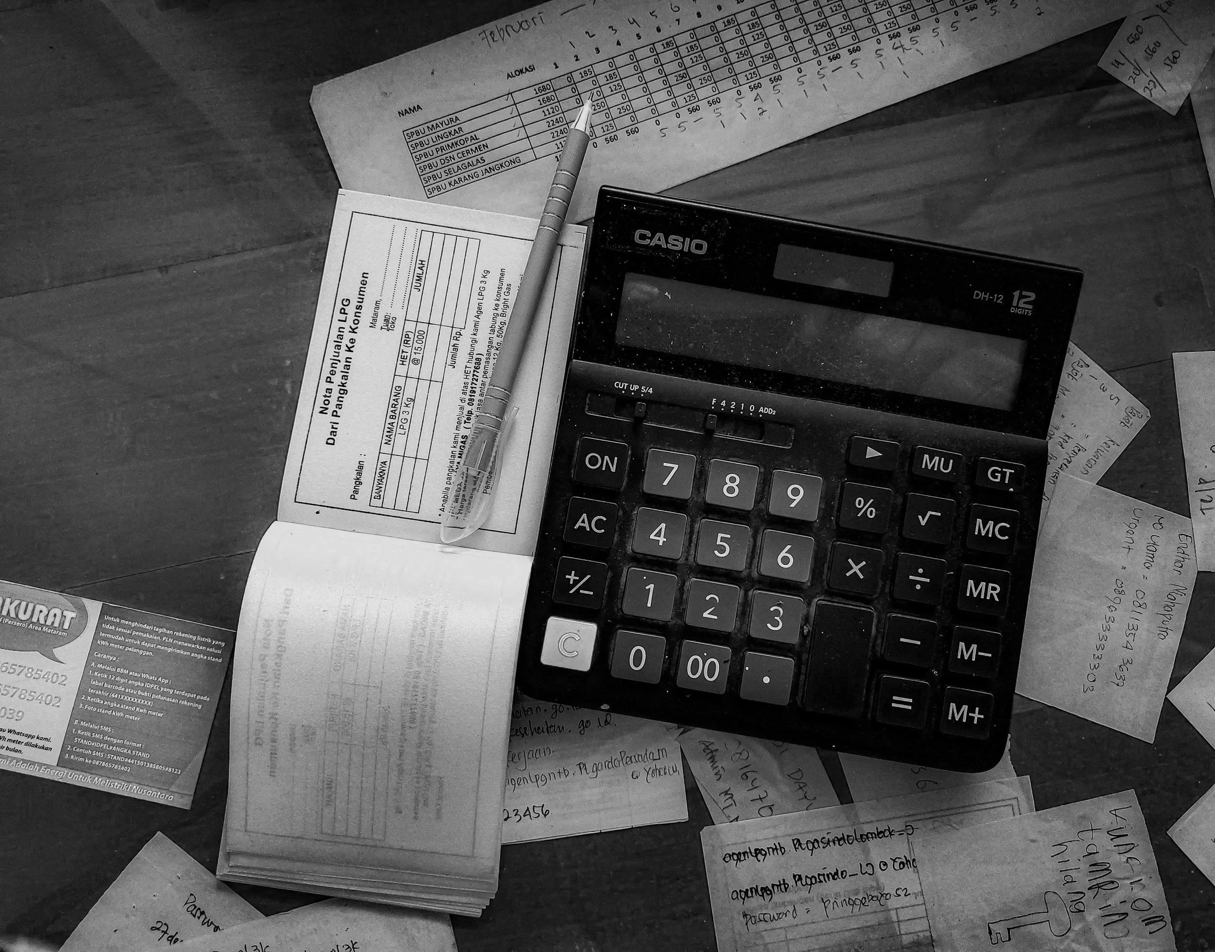 A black and white photograph showing scattered papers, a pen, a calculator, and a list of items, possibly receipts or notes, on a wooden surface.