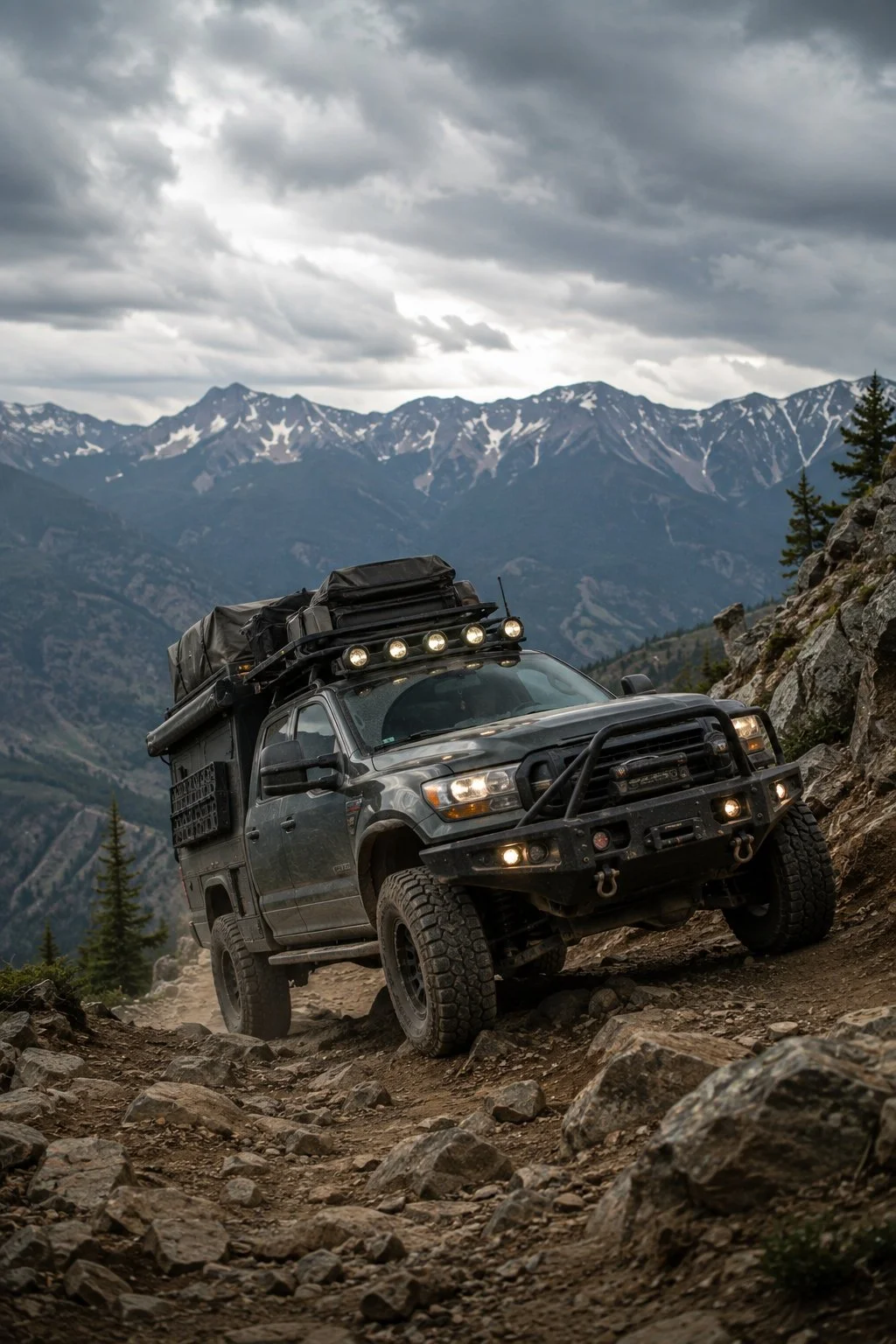 A rugged off-road vehicle driving on a rocky mountain trail with snow-capped mountains and cloudy sky in the background.