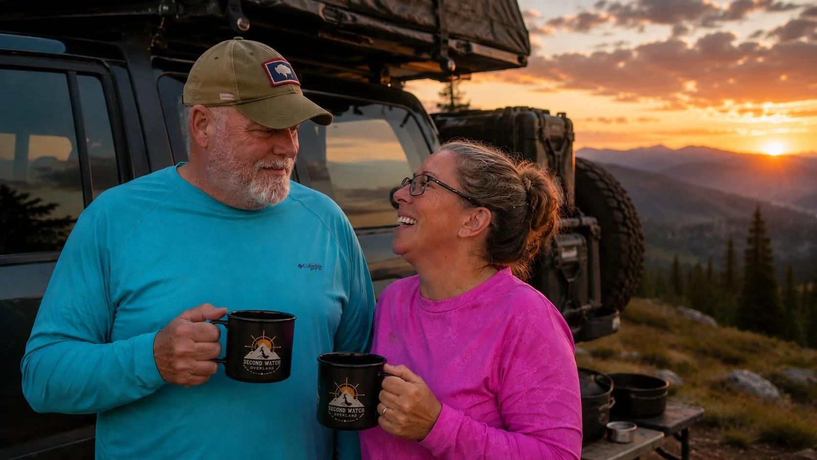 An older man and woman smiling and holding black mugs with a mountain logo, standing outdoors near a vehicle at sunset with a scenic mountain view in the background.