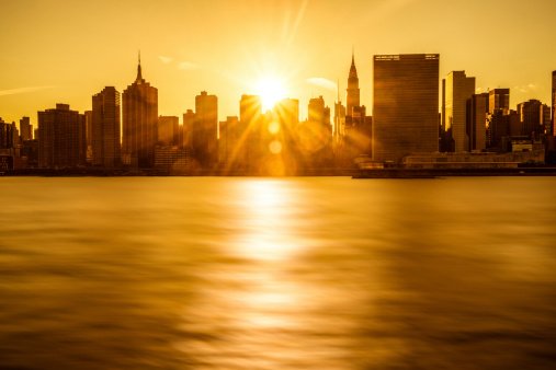 Sunset over a city skyline with tall buildings and water in the foreground.