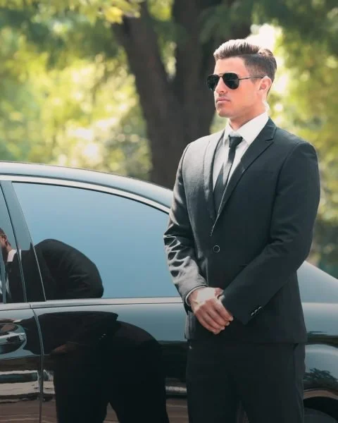 Man in a black suit and sunglasses standing outdoors near a black car, with trees and greenery in the background.