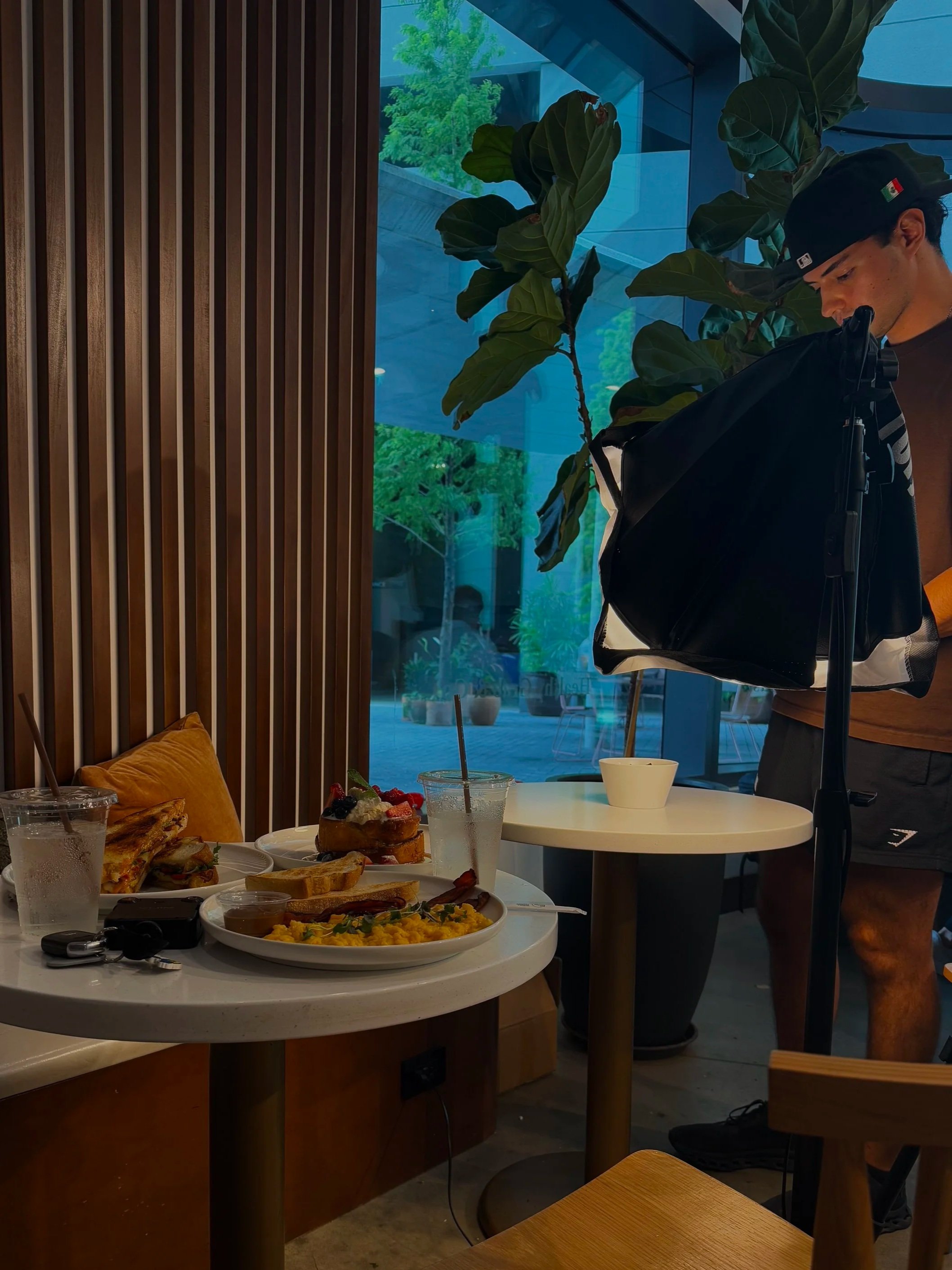 A table with various foods including a plate of pancakes with berries, a bowl of scrambled eggs, slices of toast, and drinks with straws. In the background, a person wearing a black cap looks at a black bag, with a large plant and large window showing greenery outside.