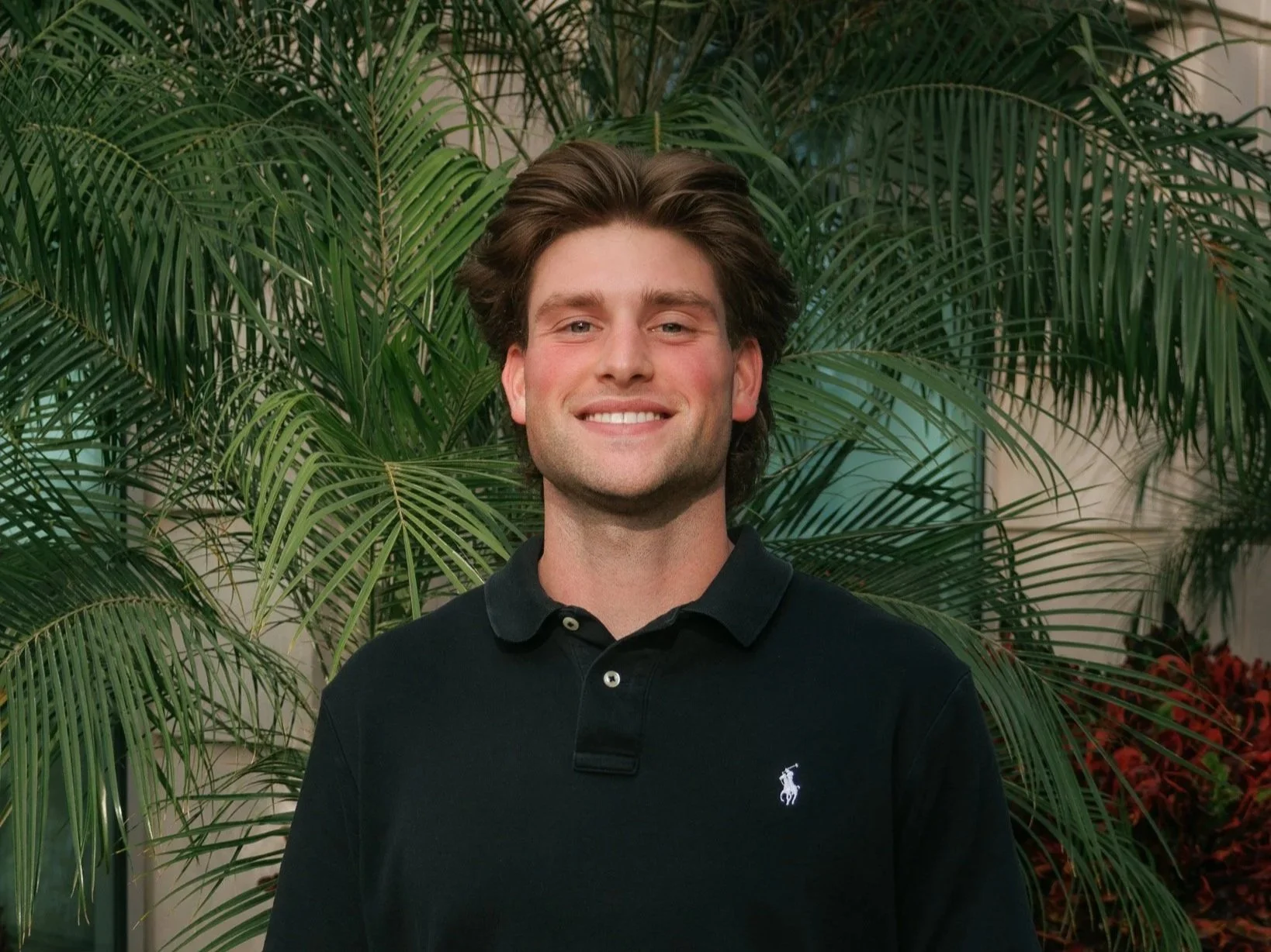 A young man with brown hair and a smile, wearing a black polo shirt with a white logo, standing in front of green palm leaves.