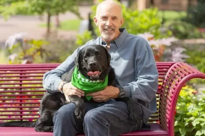 An older man with a beard sitting on a red park bench holding a black Labrador dog wearing a green bandana. The man is smiling and looking at the camera, and the background features greenery and a blurred outdoor park scene.