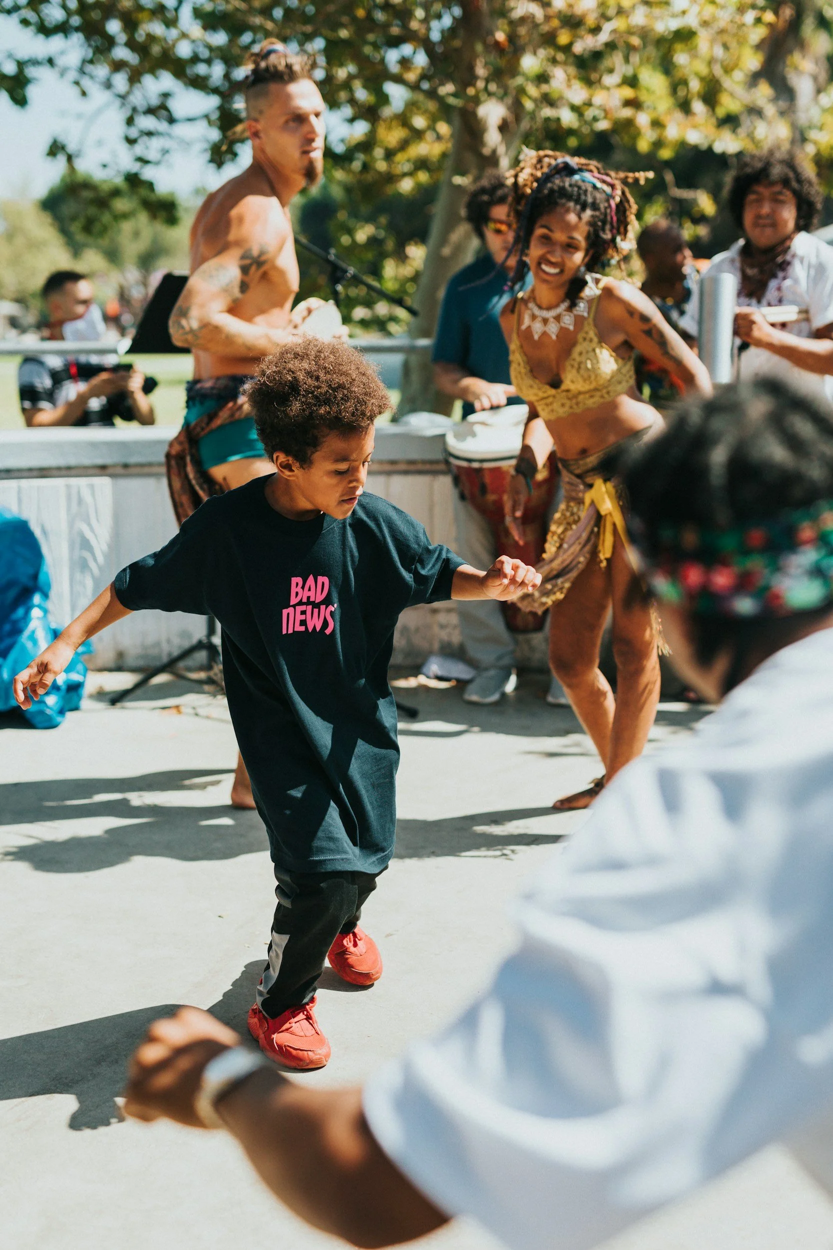 Young boy wearing a black t-shirt with pink text, dancing outdoors during a sunny day with people and trees in the background.
