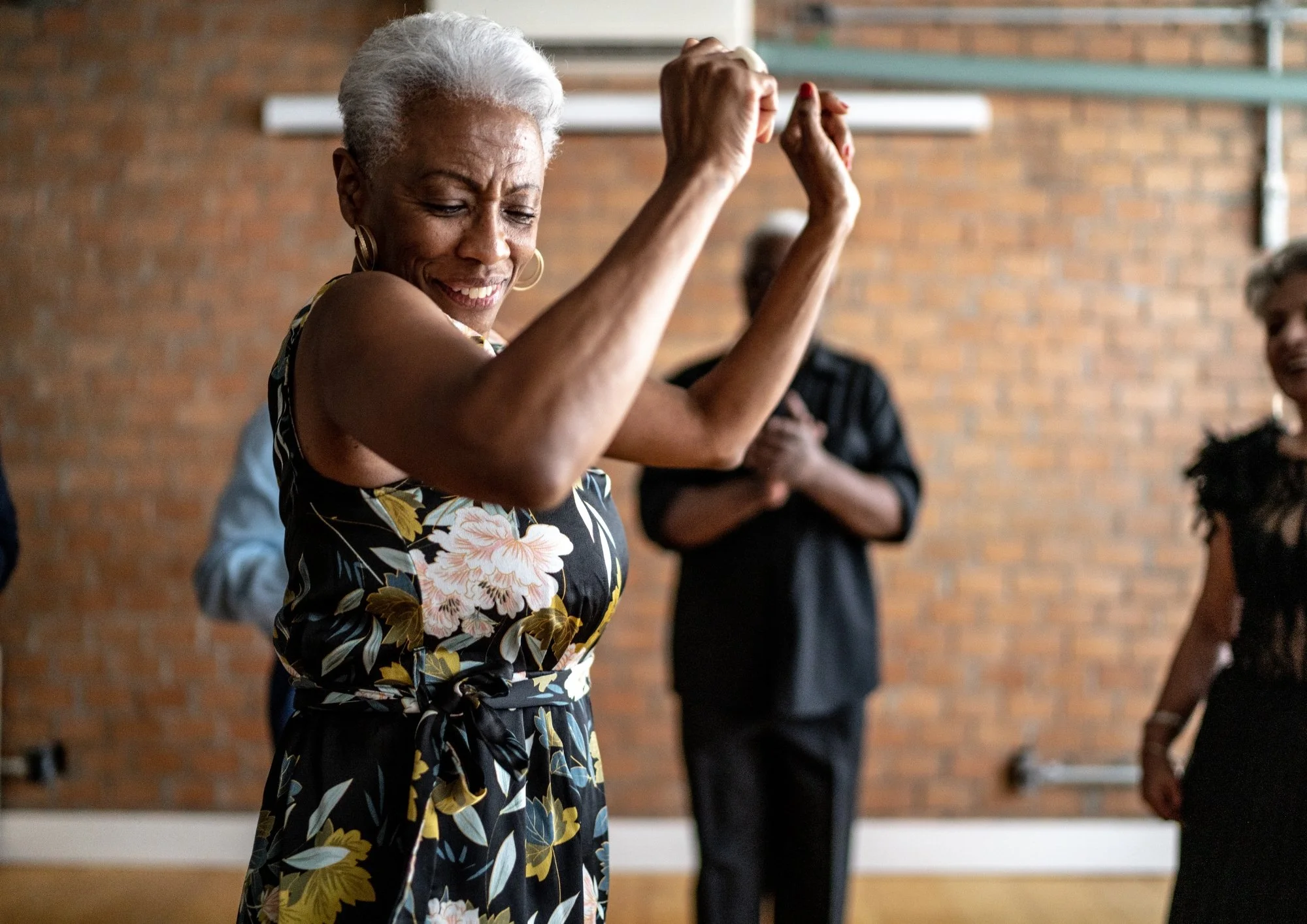 An older woman with gray hair, wearing a floral dress, dancing with her eyes closed in a community or dance class, with a brick wall background.