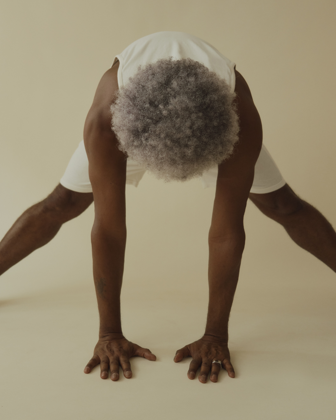 An elderly man with grey hair doing a downward facing dog yoga pose against a plain beige background.