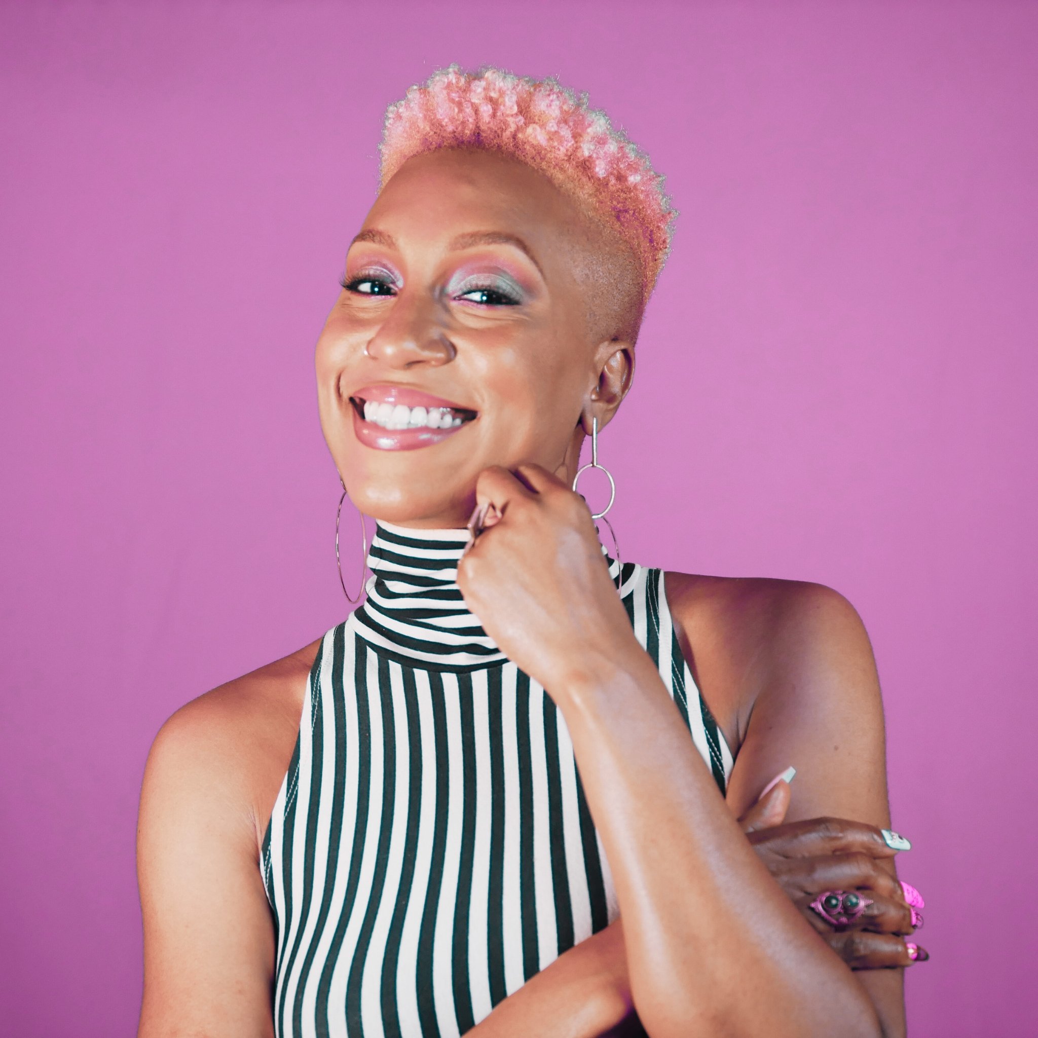A woman with short, curvy hair dyed pink and blonde, smiling against a purple background. She is wearing a striped tank top, hoop earrings, and has colorful jewelry on her wrist.