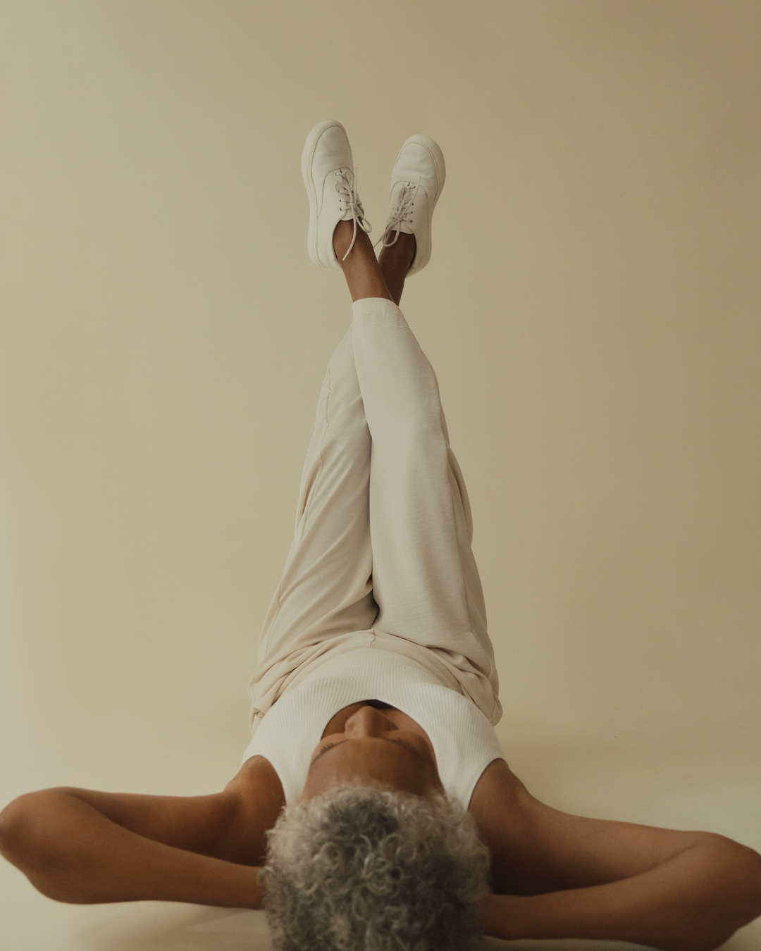 An elderly woman with curly gray hair lying on her back with her legs raised and crossed, wearing a white sleeveless top, beige pants, and white sneakers, against a plain beige background.
