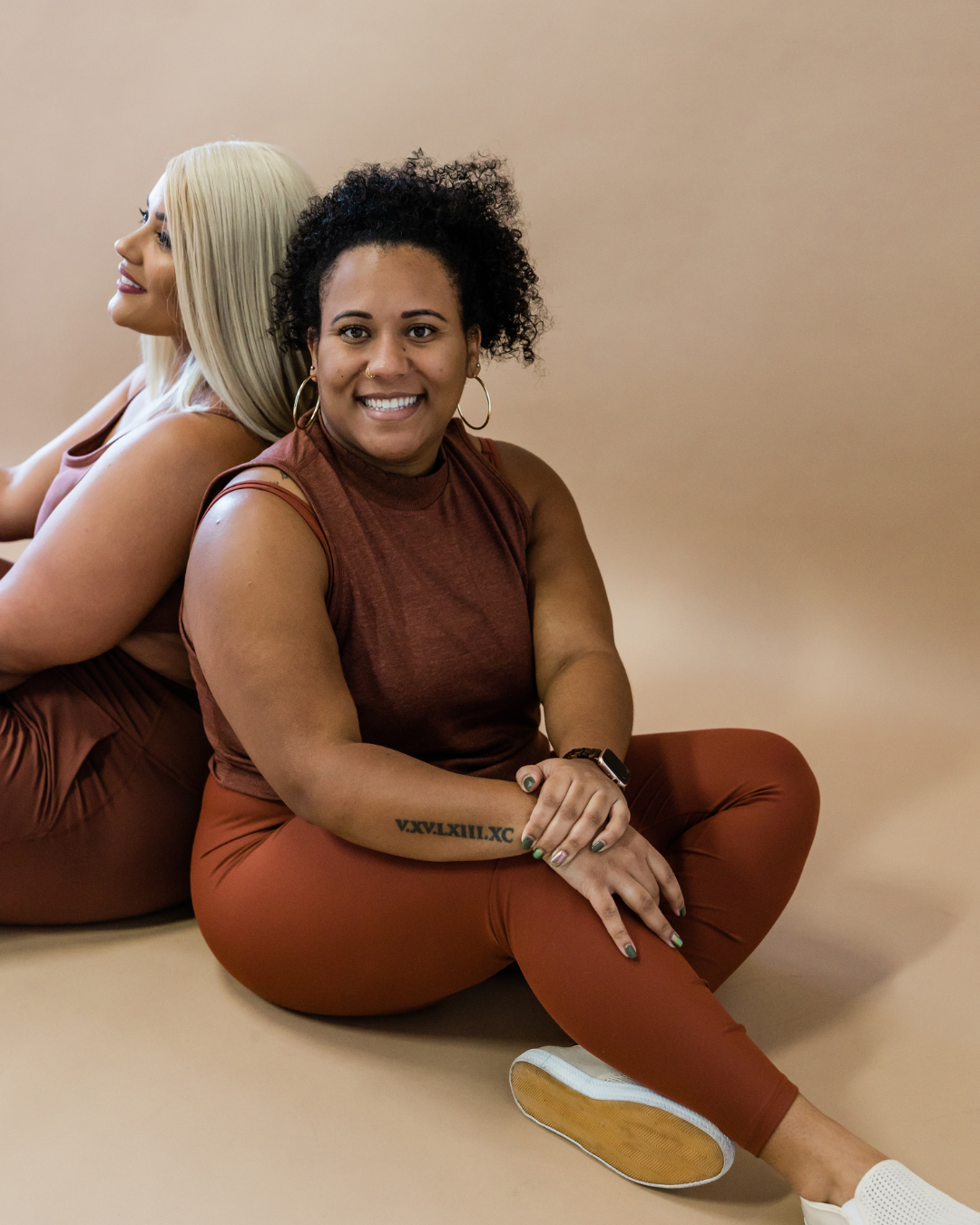 Two women sitting back-to-back on the floor, smiling at the camera, with a plain beige background. The woman in front has curly hair, hoop earrings, and a tattoo on her forearm, and is wearing a sleeveless brown top and matching leggings.