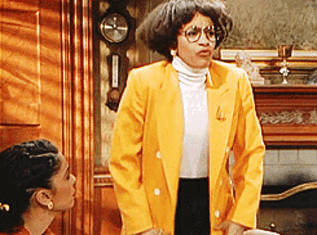A woman with curly hair, glasses, and wearing a yellow blazer standing indoors, with another woman sitting and looking at her.