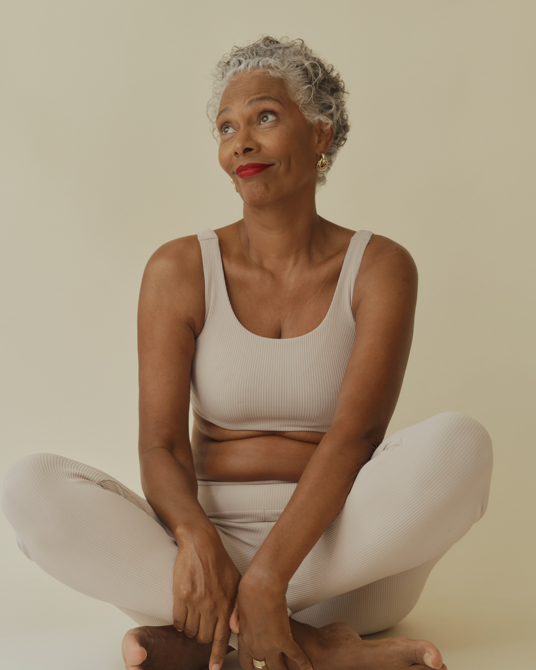 An older woman with short gray curly hair, wearing a beige sports bra and matching leggings, sitting cross-legged on the floor with a playful expression and red lipstick.