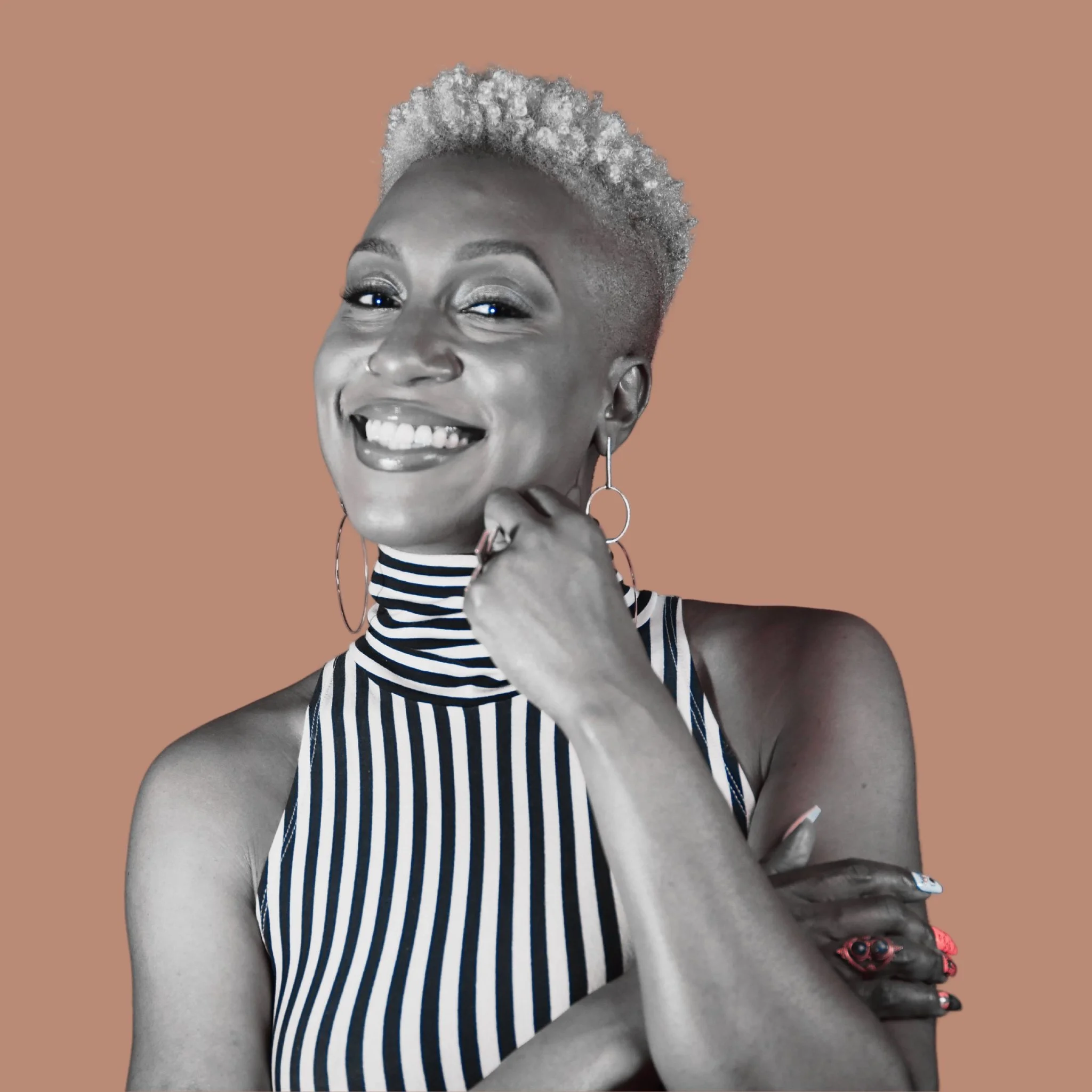 A black and white portrait of a woman with a short, textured hairstyle, smiling and wearing a striped sleeveless top, with hoop earrings and a decorated skirt, set against a plain background.