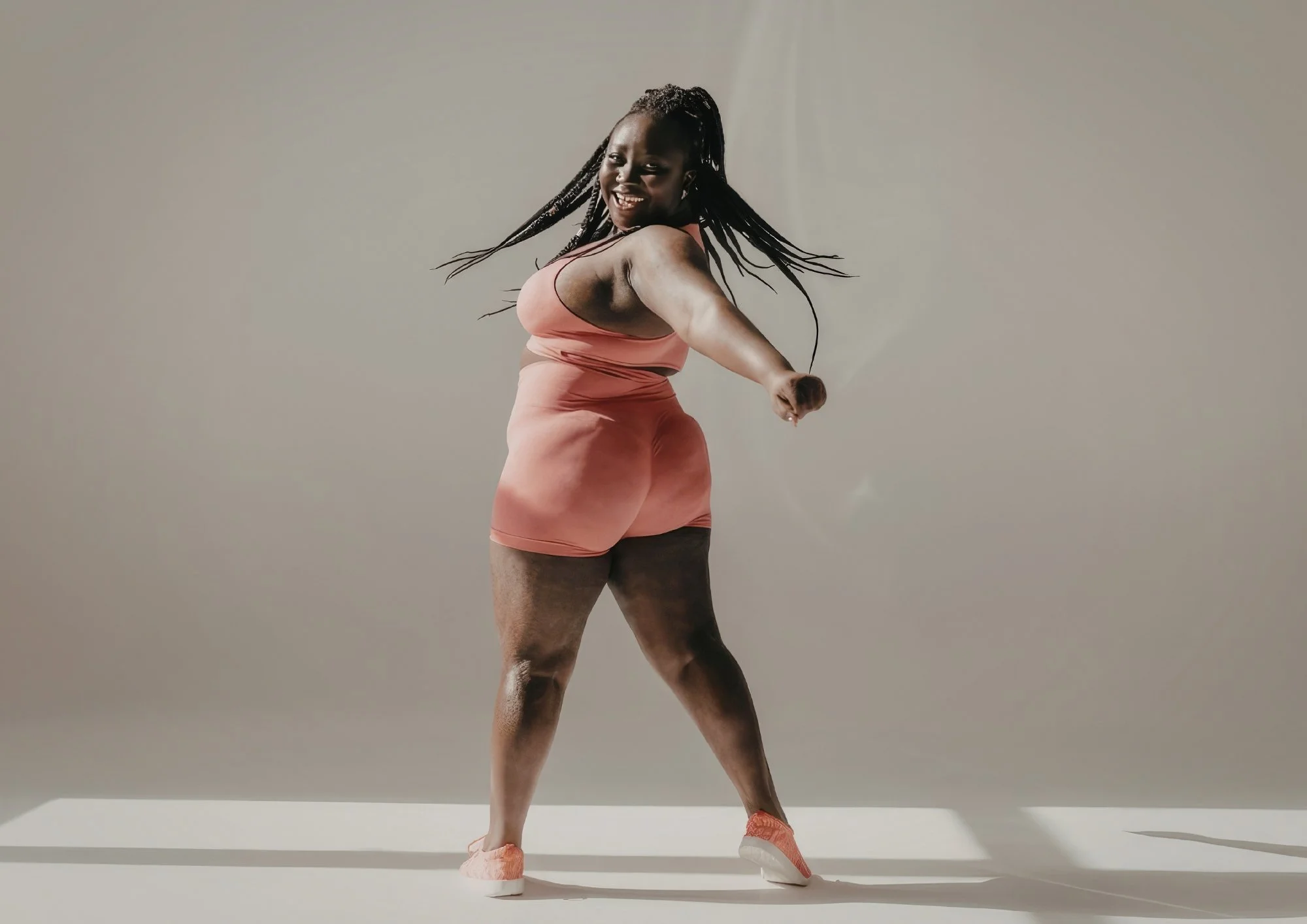 A woman dancing in a peach-colored dress and sneakers, smiling, against a plain gray background.