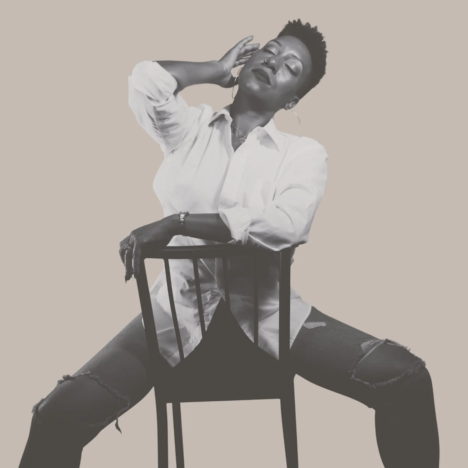 A woman with short curly hair and hoop earrings, wearing a white shirt and ripped jeans, sitting on a chair with her legs spread wide. She has her eyes closed and one hand touching her face, with a calm and relaxed expression against a plain background.
