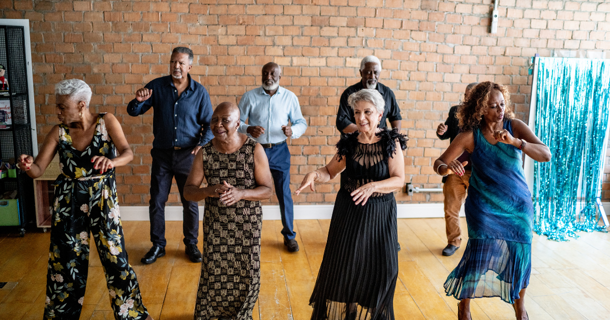 Six diverse adults dancing together in a room with a brick wall and wooden floor, celebrating at a social gathering.