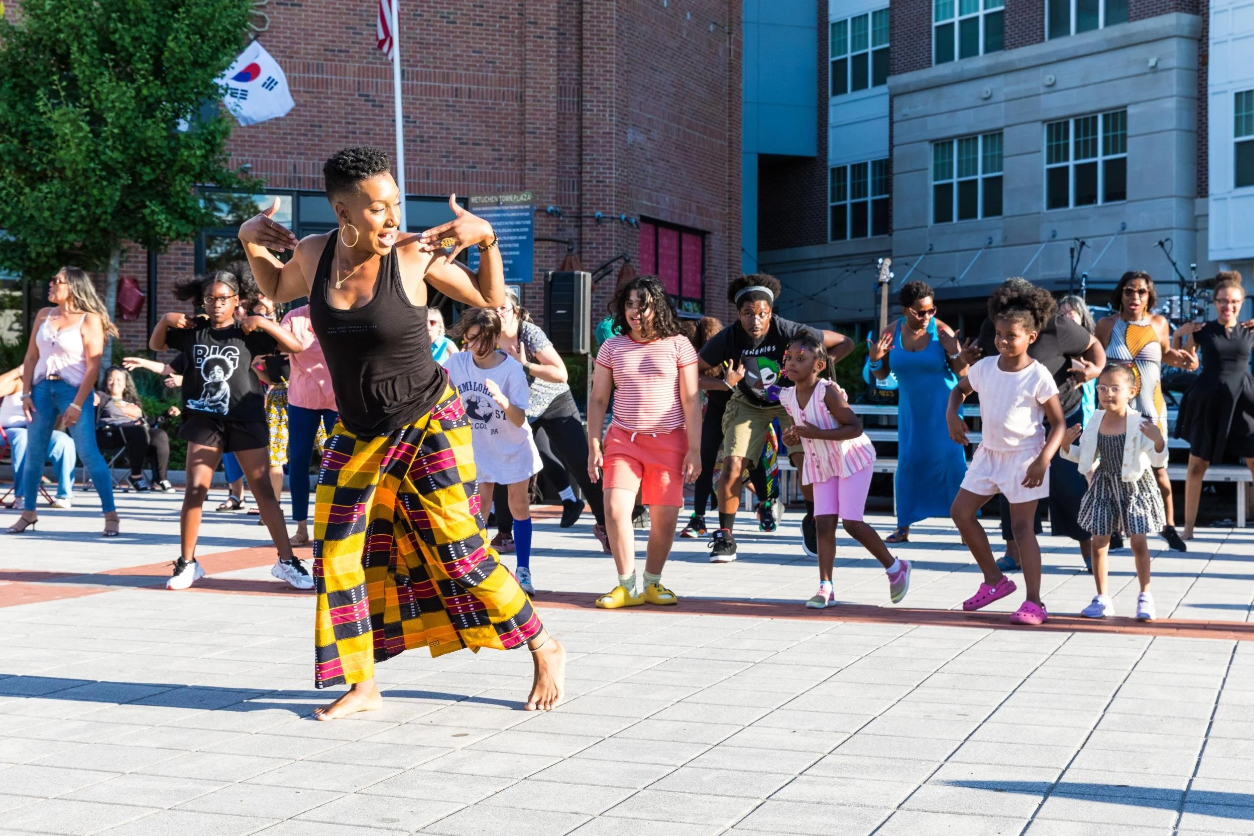 A woman dancing barefoot in front of a group of children and adults on an outdoor city plaza during daytime.