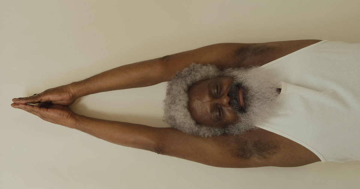 An elderly man with gray beard and hair practicing yoga, stretching with arms extended overhead, wearing a white tank top.