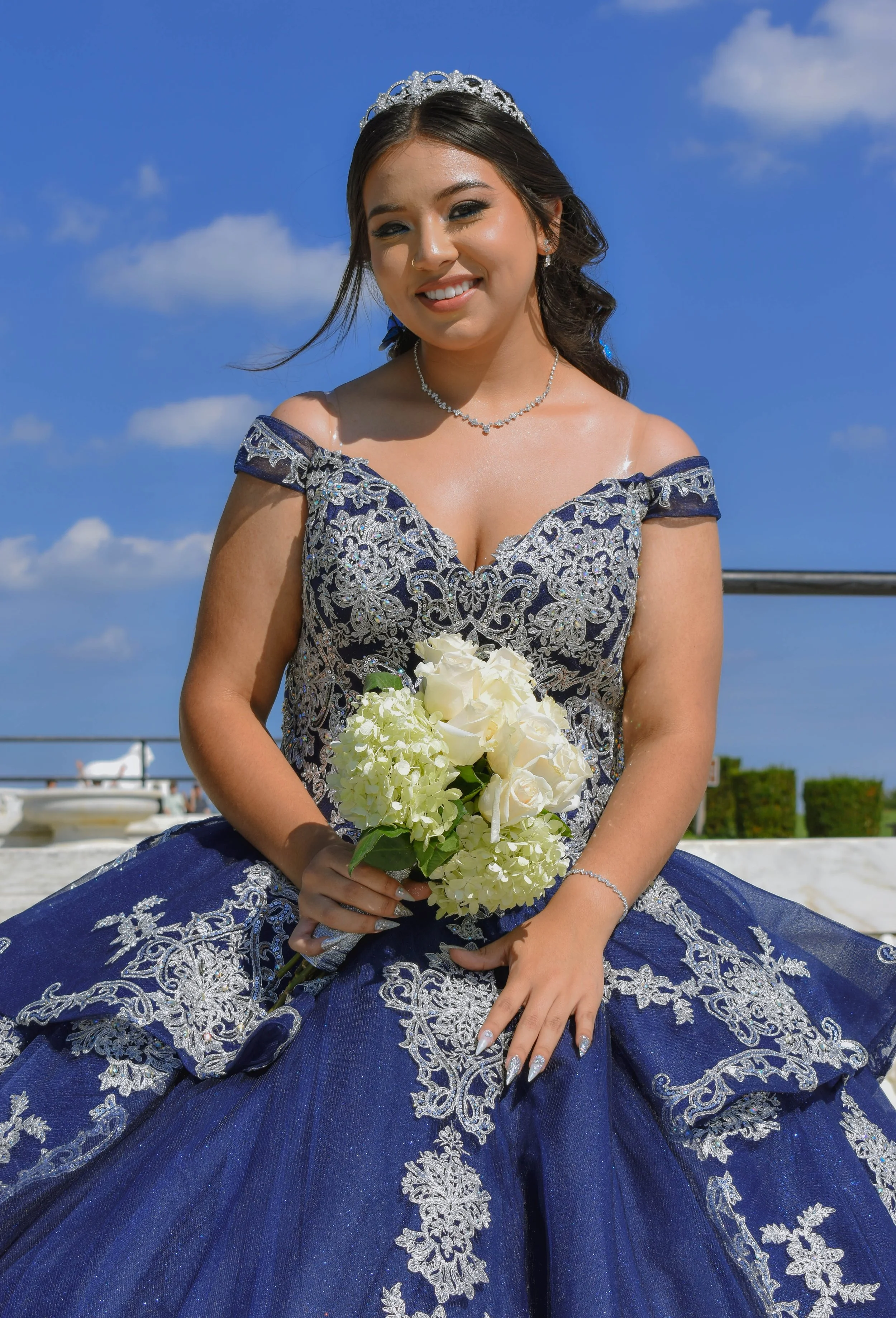A young woman in a royal blue and silver embroidered gown, wearing a tiara and necklace, sitting outdoors with a bouquet of white roses, smiling at the camera against a blue sky with a few clouds.