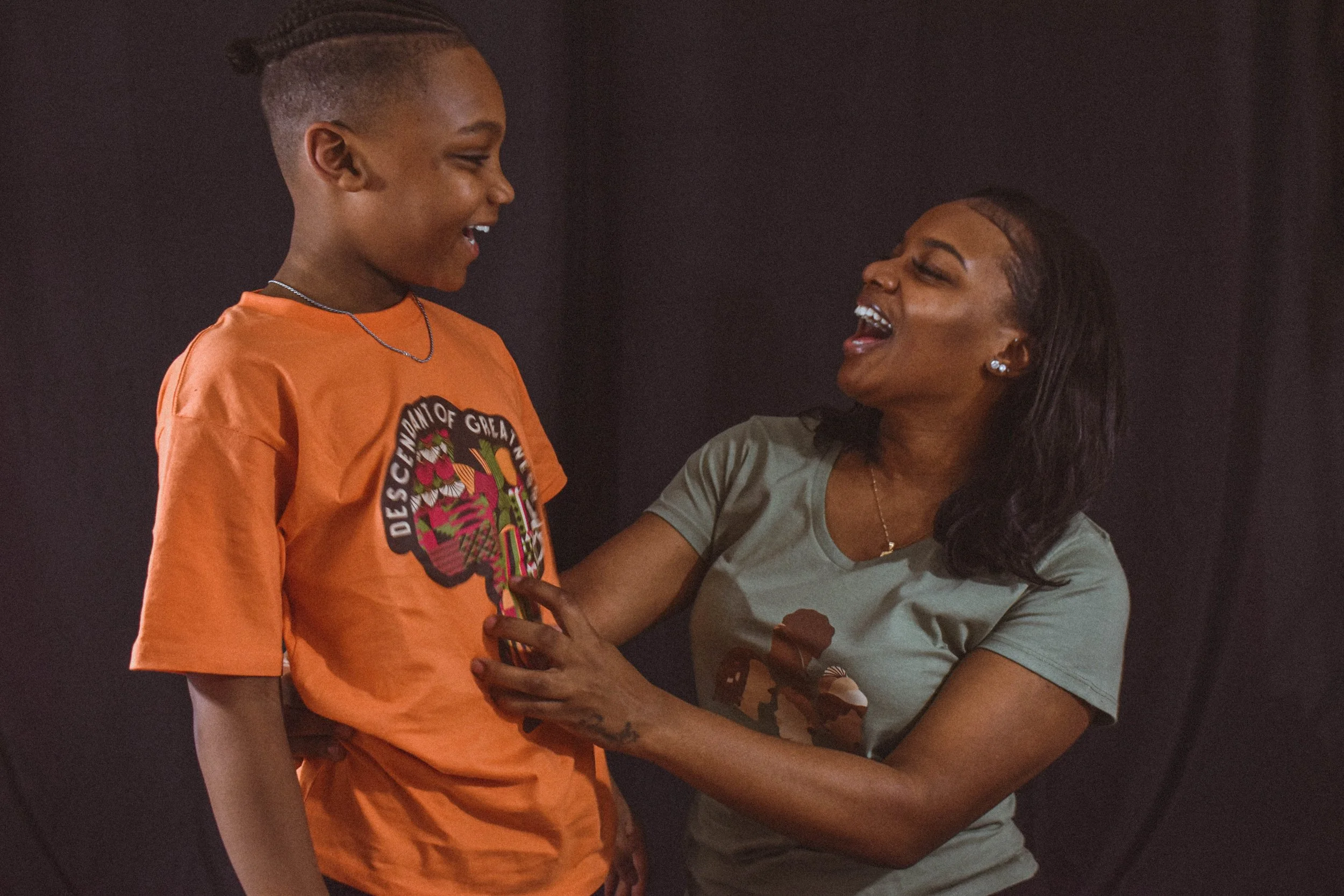 A young boy wearing an orange shirt and a woman wearing a gray shirt sharing a joyful moment, looking at each other and smiling against a dark background.