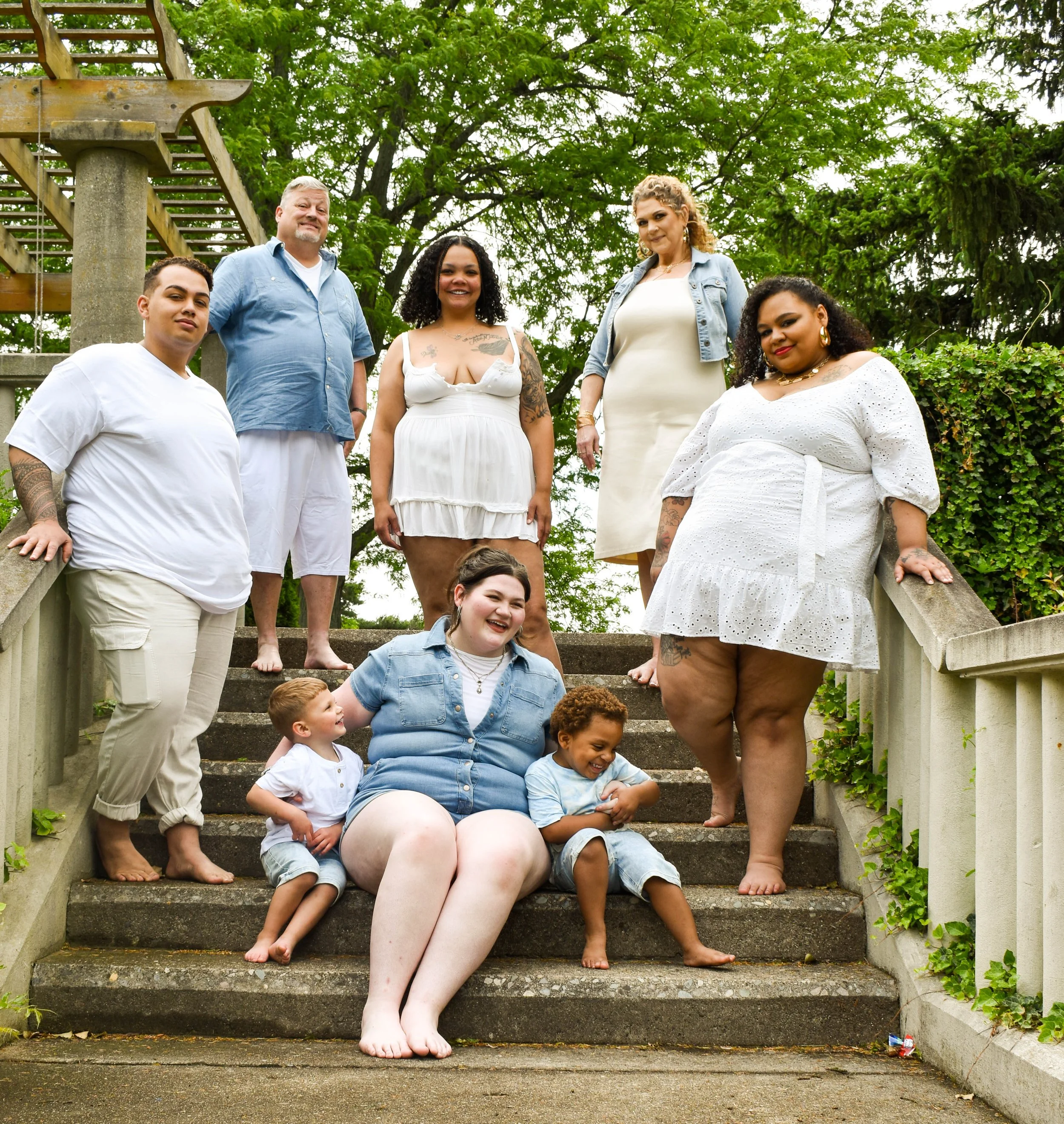 A group of people gathered on outdoor steps surrounded by greenery. The group includes adults and children, with a focus on multicultural diversity. Some individuals are smiling, and one woman is leaning on the handrail, showcasing a casual and friendly atmosphere.