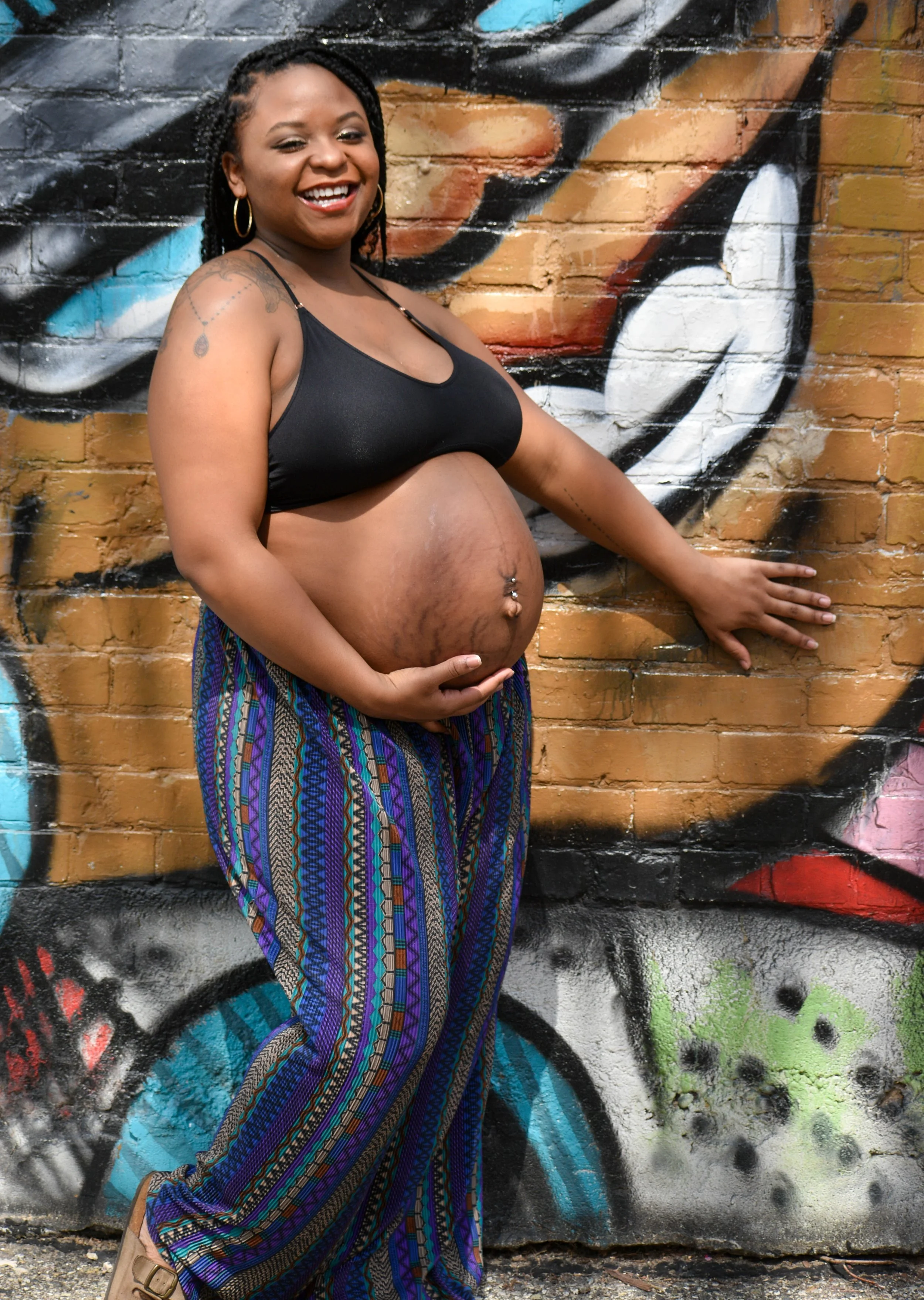 A smiling woman with dreadlocks and hoop earrings, wearing a black crop top and colorful, patterned pants, standing in front of a graffiti-covered brick wall.