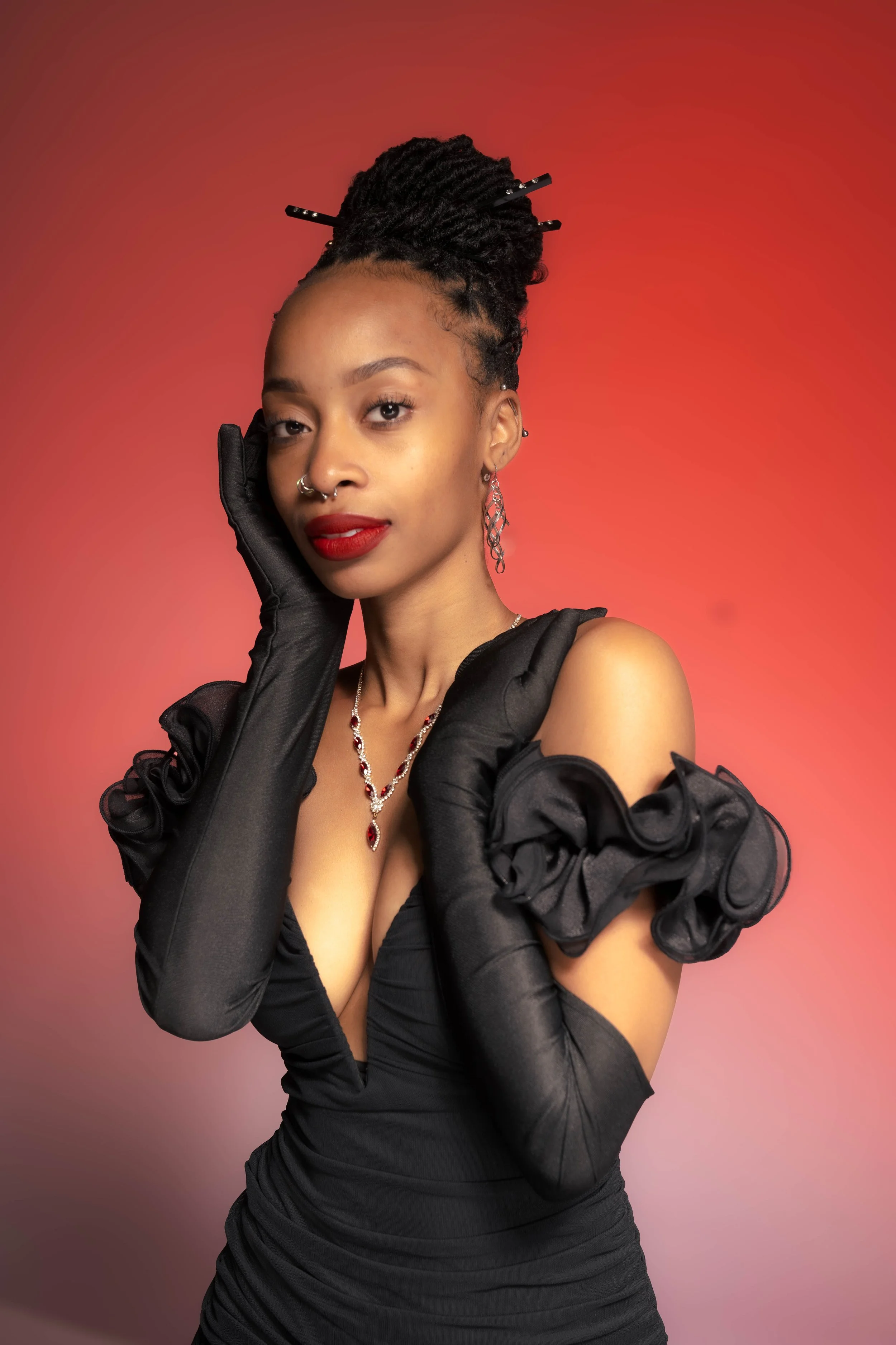A woman with dark curly hair styled in a high updo, wearing red lipstick, a black dress with ruffled sleeves, long black gloves, and jewelry including a necklace, earrings, and a septum piercing, holding one hand to her face against a gradient red background.