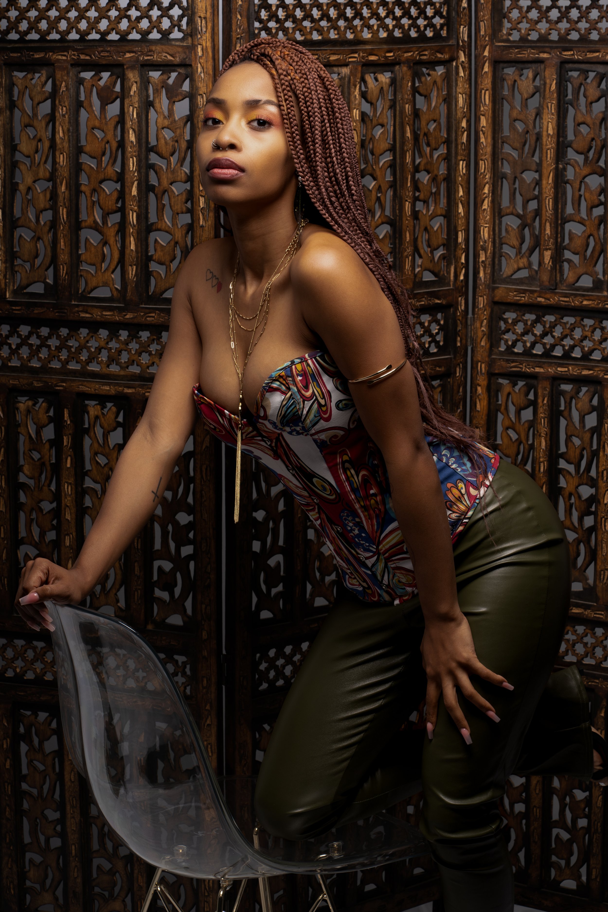 A woman with braids, tattoos, and gold jewelry poses in front of a wooden lattice screen, wearing colorful top and leather pants, with a transparent motorcycle helmet positioned at the bottom left.