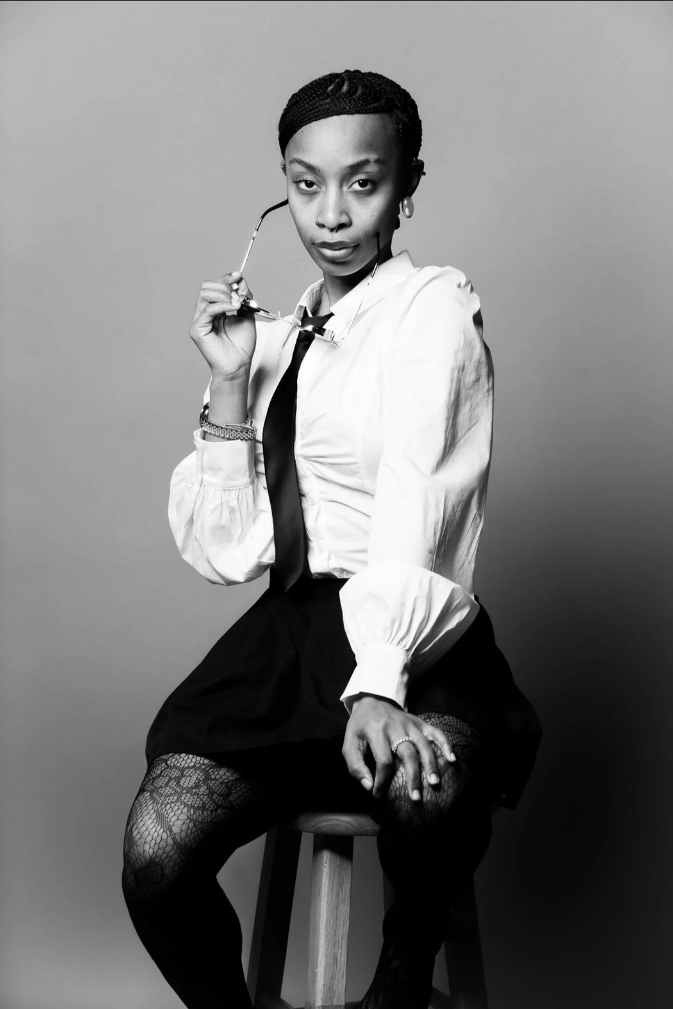 Black and white portrait of a woman sitting on a stool, holding glasses in her right hand, wearing a white blouse, a black tie, fishnet stockings, and lace gloves, with short braided hair, against a plain background.