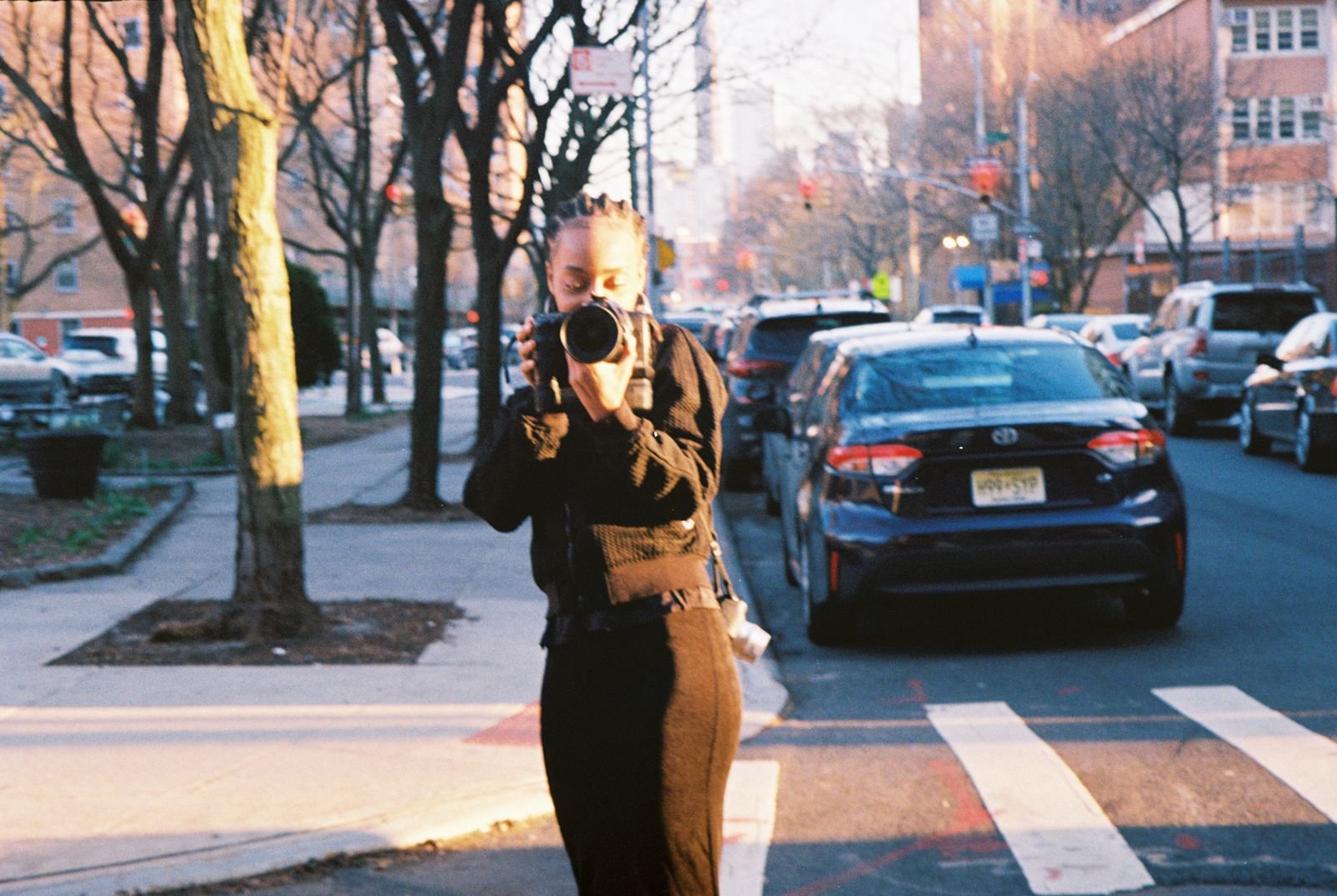 A person holding a camera takes a photo in an urban street setting with trees, parked cars, and buildings in the background, during sunset or golden hour.