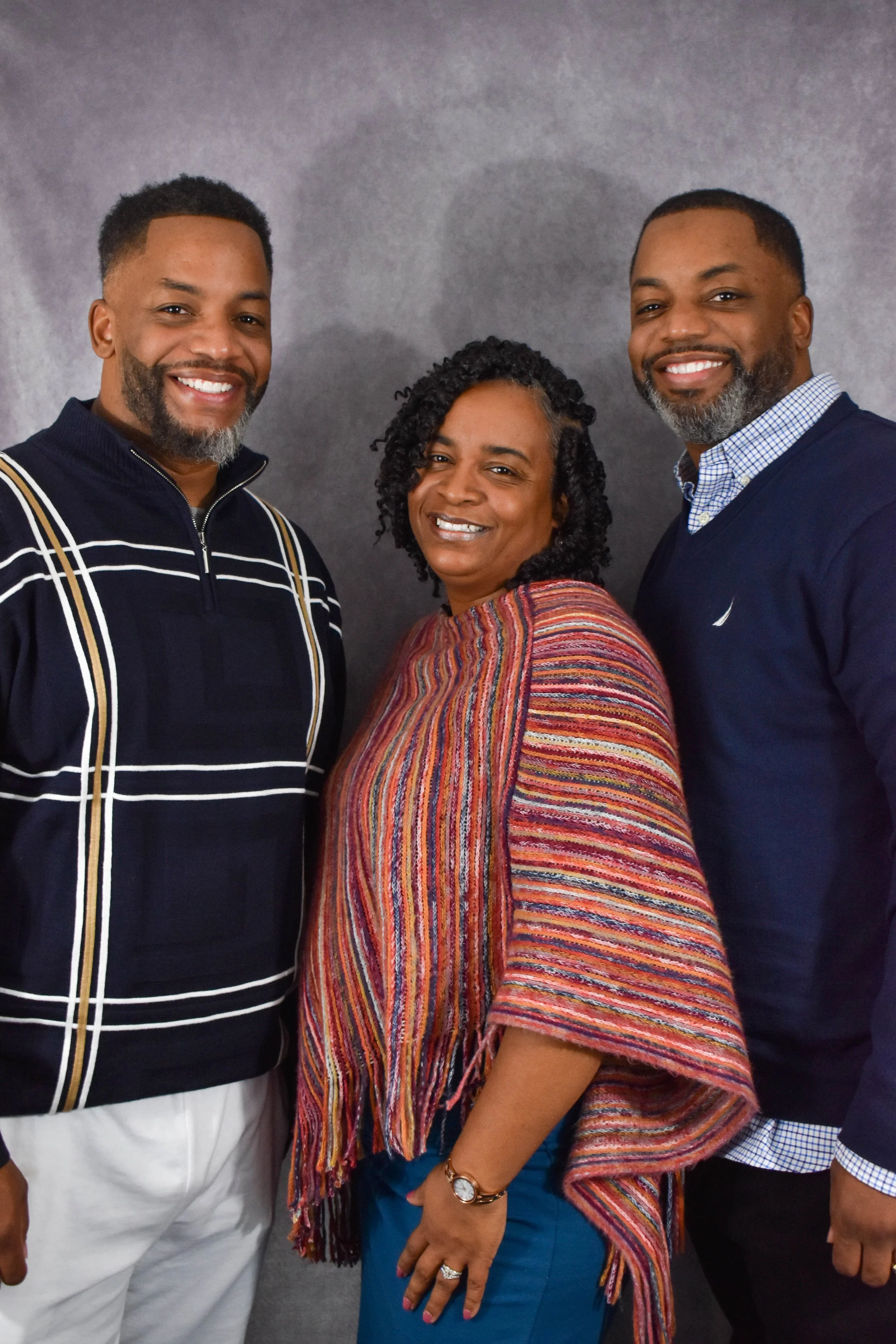 A group of three smiling adults standing close together against a gray background, two men and one woman, dressed casually.