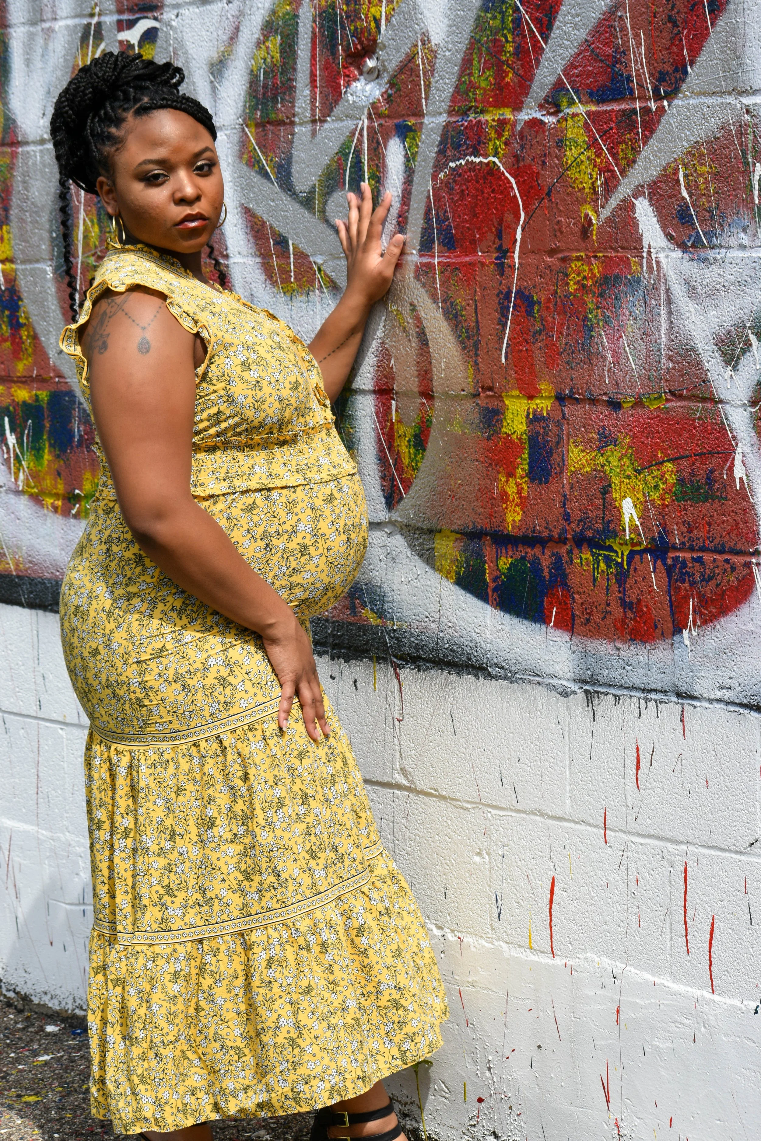 A pregnant woman in a yellow floral dress standing next to a graffiti mural on a concrete wall.