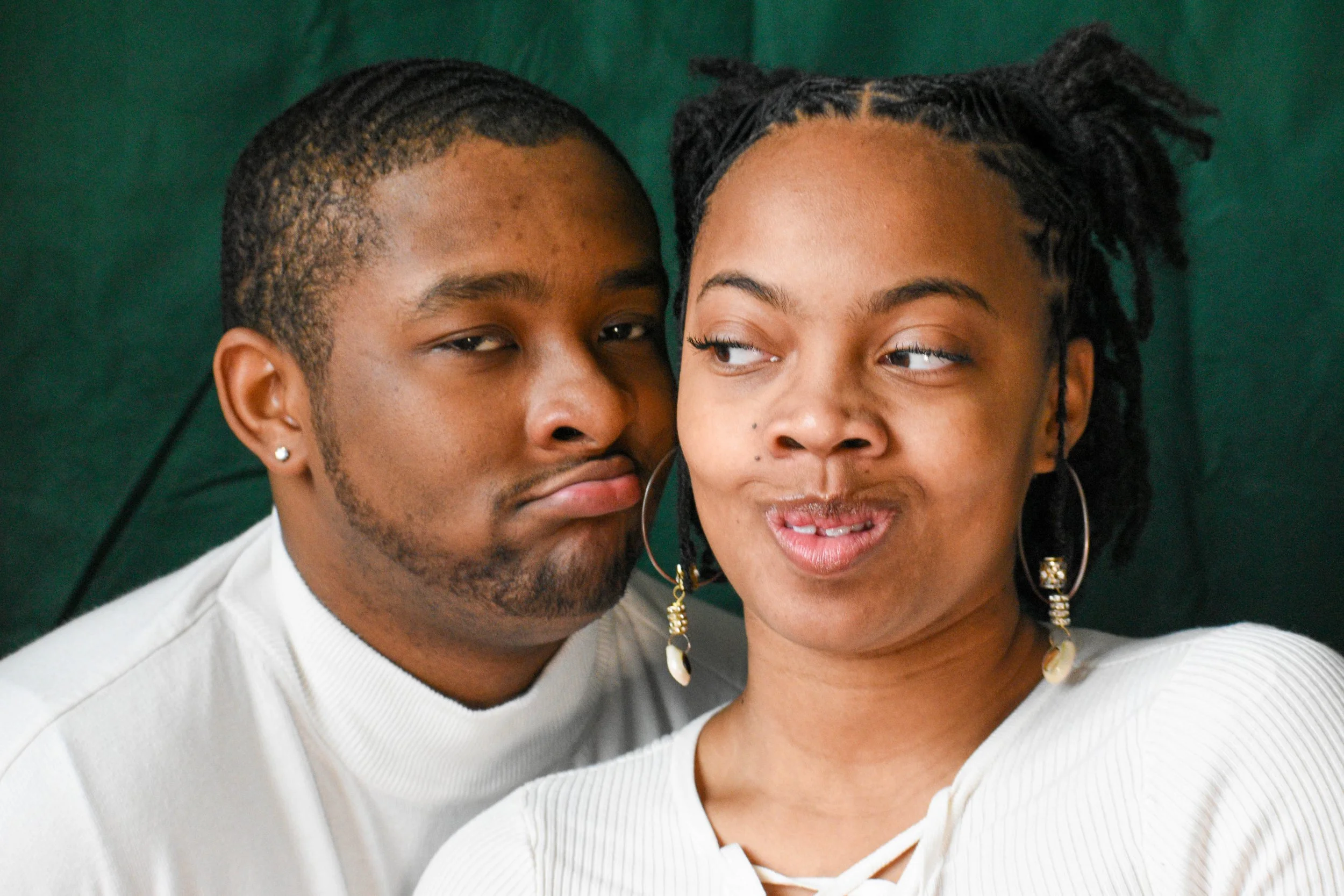 Close-up portrait of a Black man and woman, both with short curly hair, smiling and looking at each other, against a dark green background.